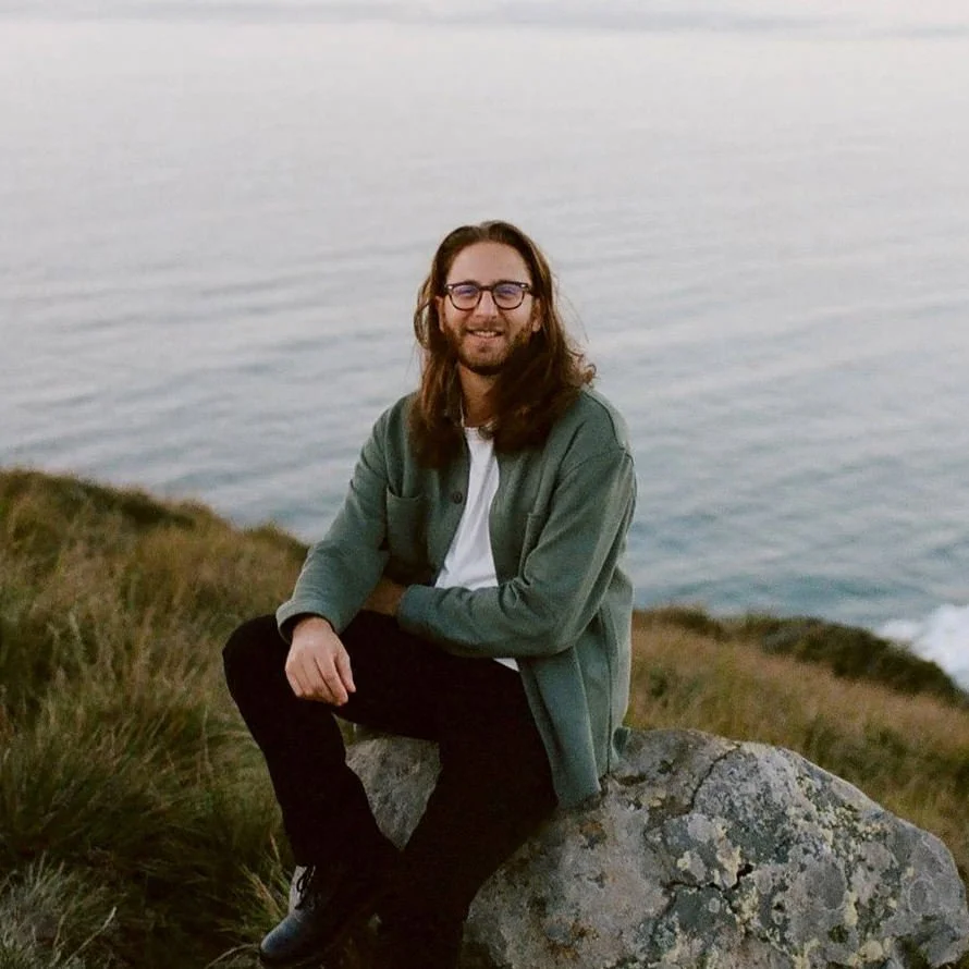 A man with long hair, glasses, and a beard sitting on a large rock near the water's edge at sunset, smiling at the camera, wearing a green jacket and black pants.