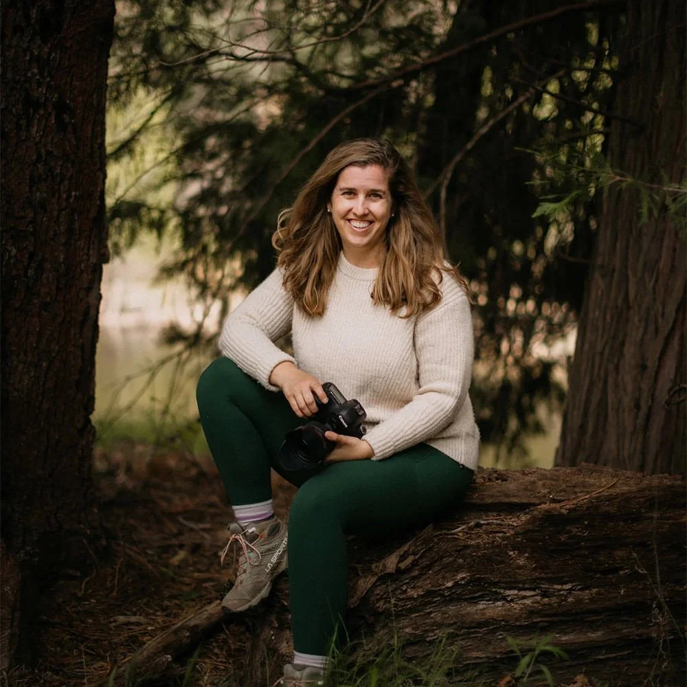 A woman sitting on a fallen log in a forest, holding a camera, smiling at the camera.