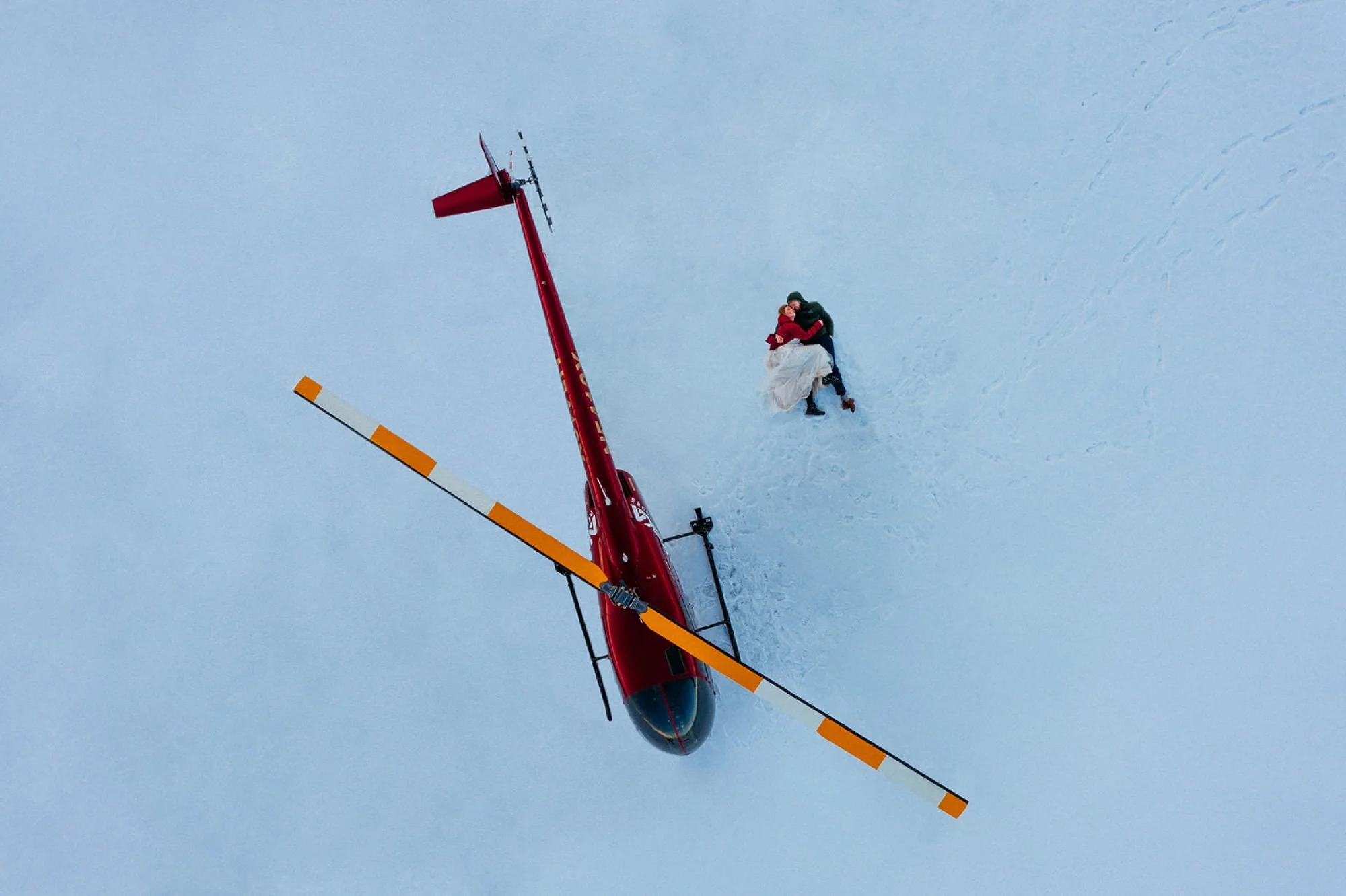 Aerial view of a red helicopter on snow with a person in wedding attire lying beside it.