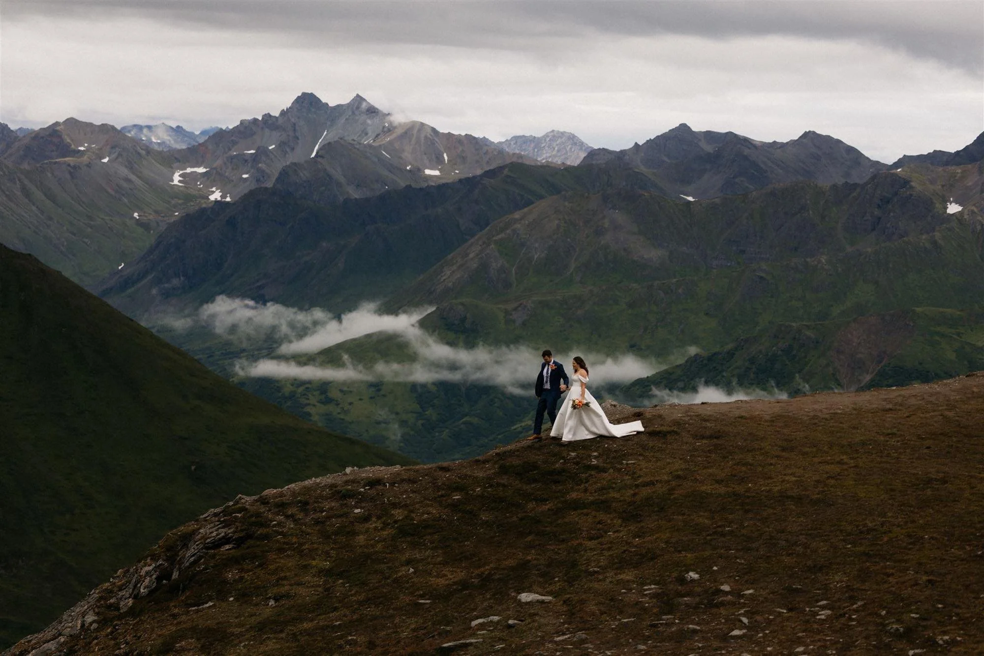 A bride and groom standing on a hillside over a mountainous landscape with clouds and peaks in the background.