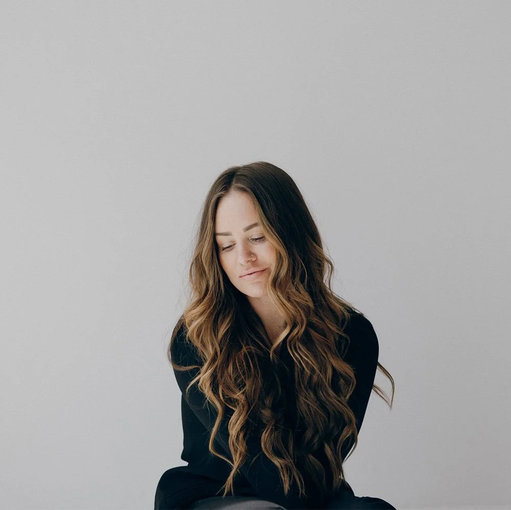 A woman with long, wavy brown hair and light skin, wearing a black top, looking downward with a soft expression against a plain light gray background.