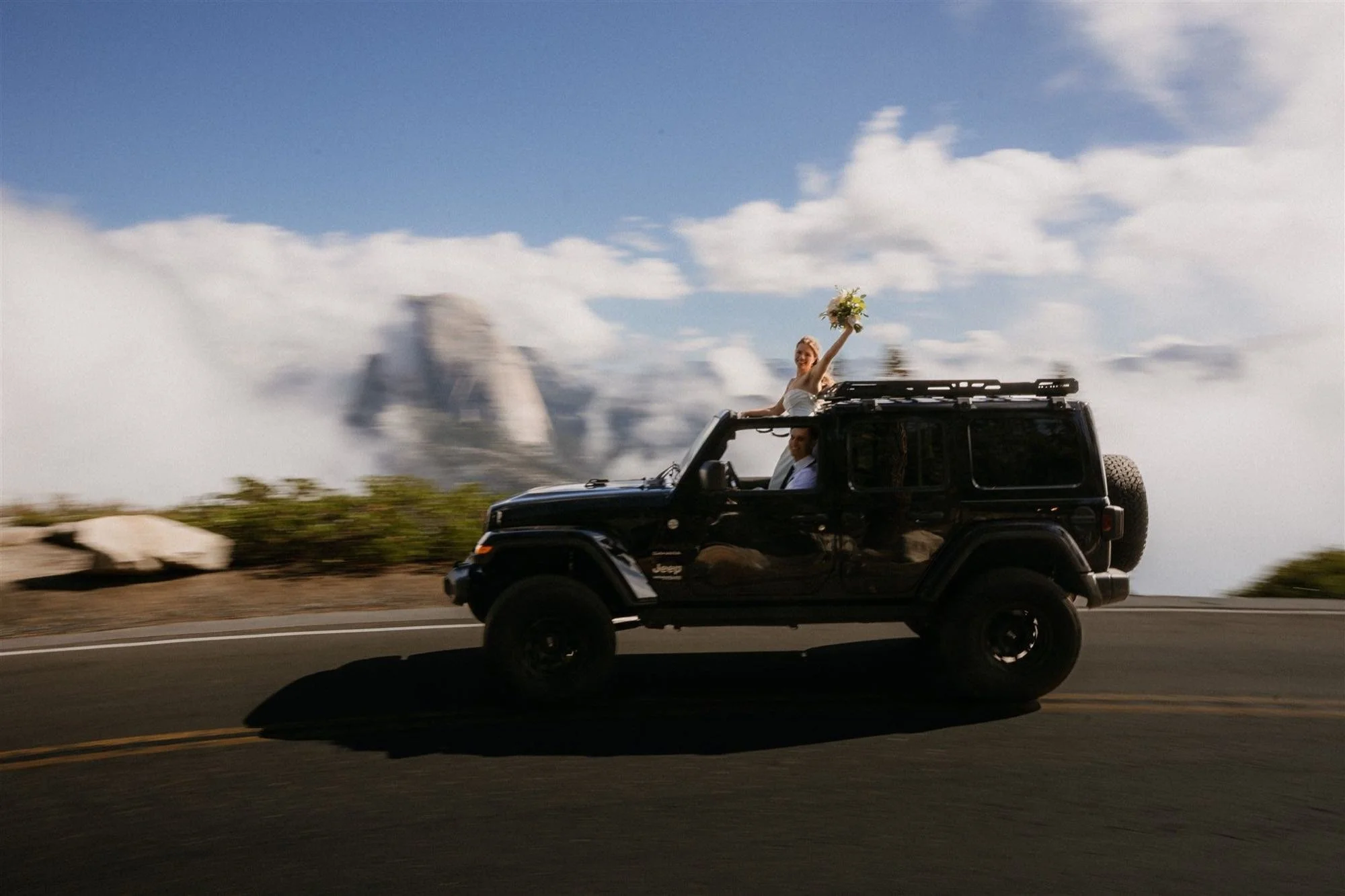 A woman in a wedding dress sitting on top of a black Jeep, holding a bouquet of flowers in the air, while the vehicle drives on a mountain road with clouds and rock formations in the background.