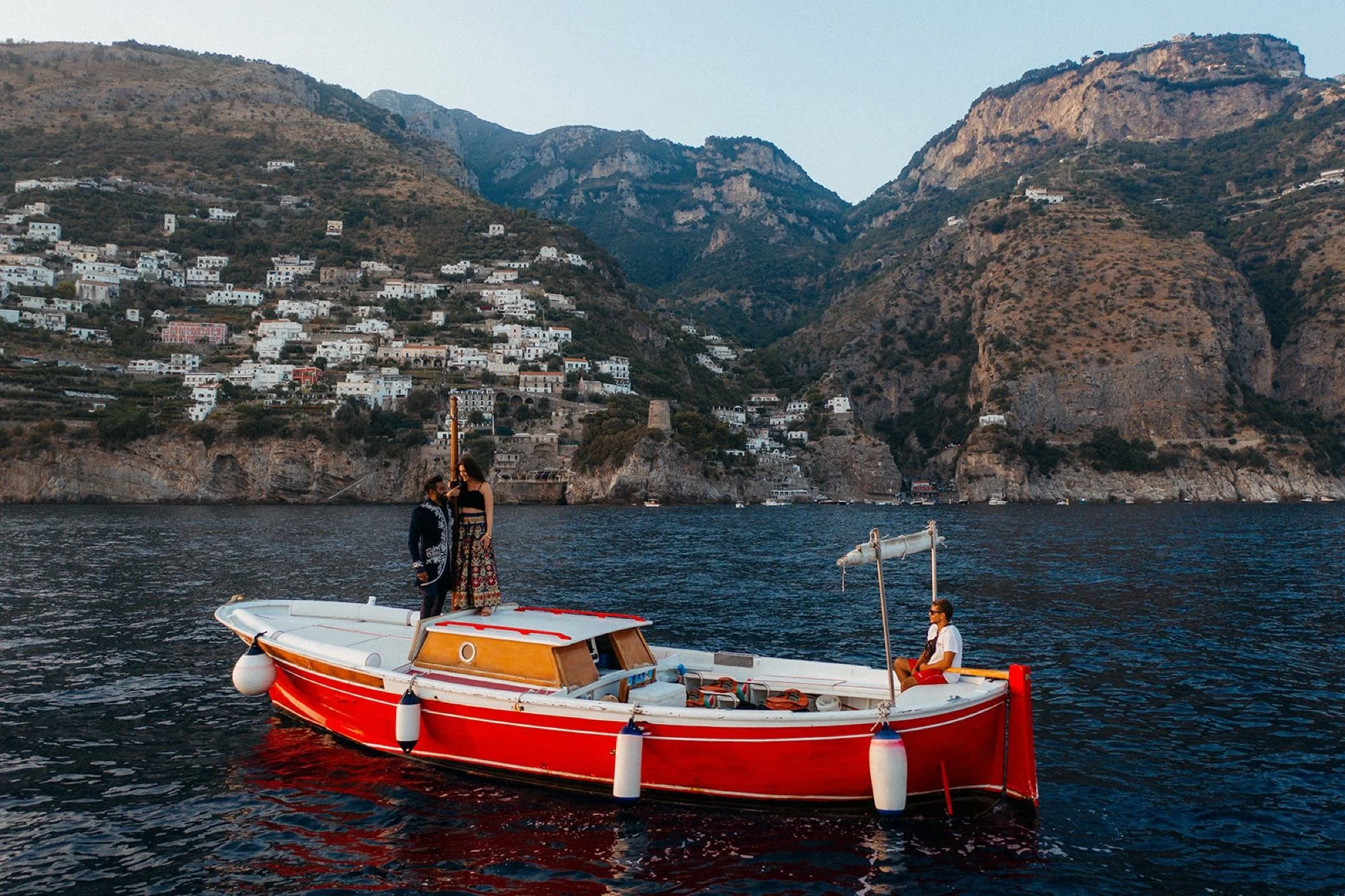 A red and white boat floating on dark water with two people standing on the boat speaking, one person sitting at the back wearing sunglasses, with mountainous terrain and white houses in the background.