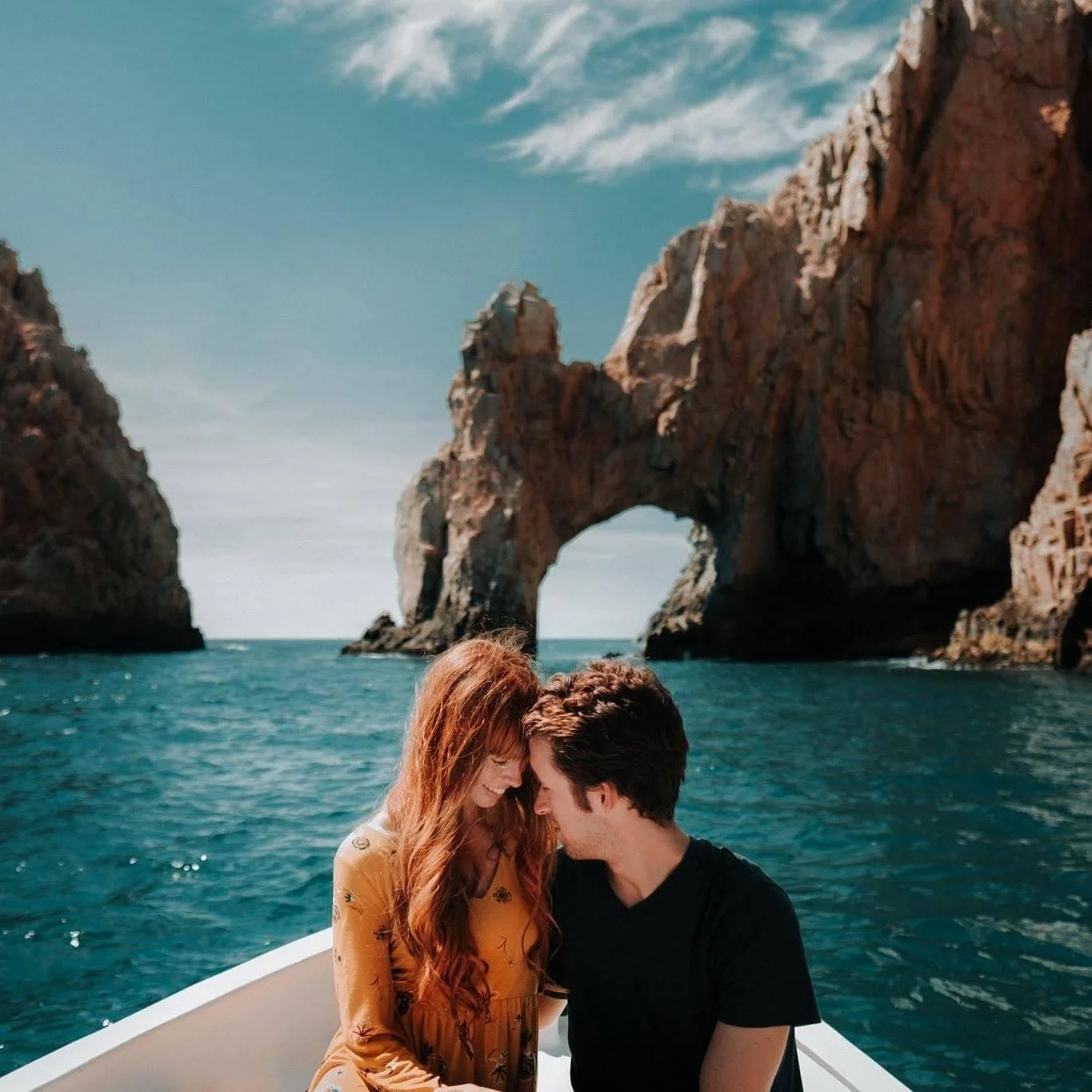 A couple sitting close together on a boat with a large natural rock arch and cliffs in the background, on a clear day with blue skies.