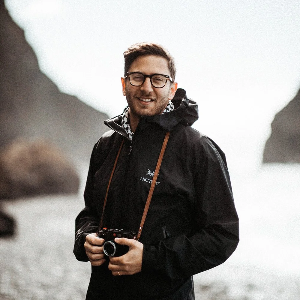 Will Khoury in a black jacket holding a camera outdoors near water and mountains.