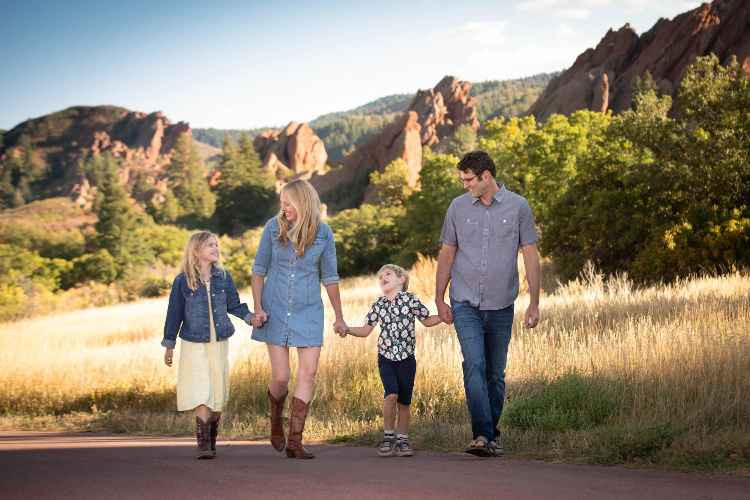 Happy family of four walking hand-in-hand on a scenic trail with red rock formations and golden grass in the background — outdoor family portrait session in Colorado by Kellie Photo, perfect for annual photo tradition.