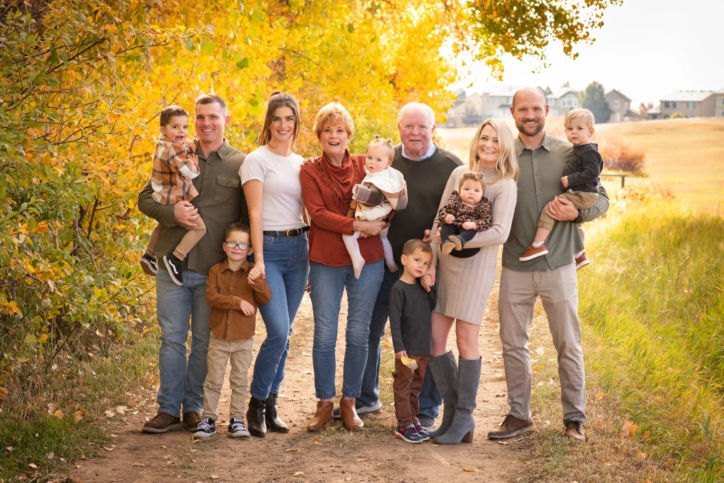 Generational portrait in a park during the fall with grandma, grandad, two dads and two moms, 4 young boys and 2 small baby girls  smiling at the camera in a family portrait session with Kellie Henriksen