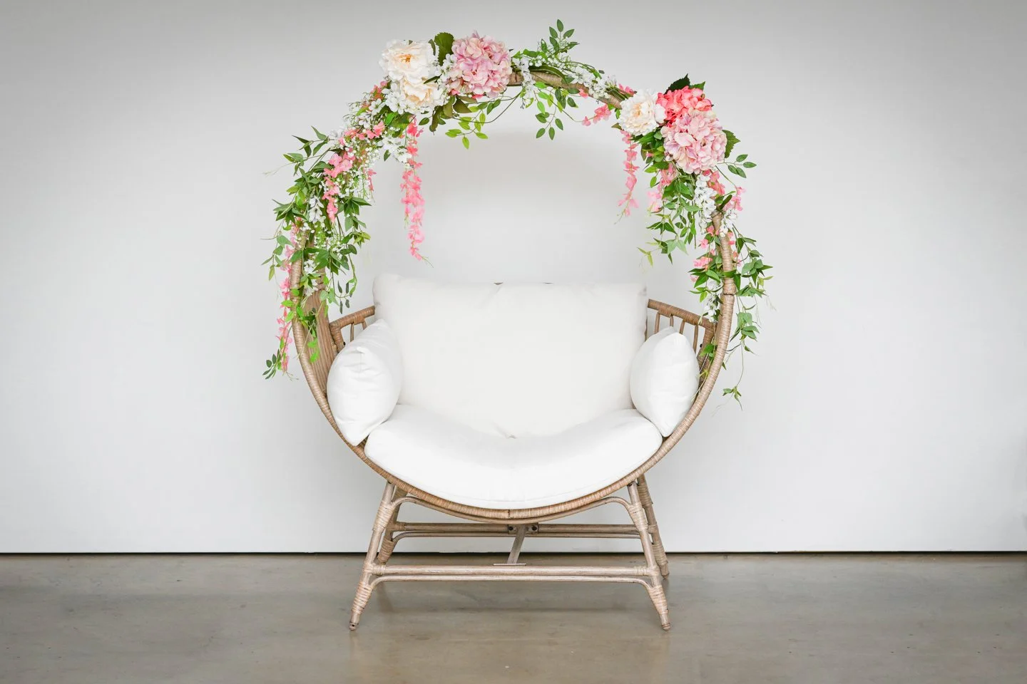 A wicker chair with white cushions and a floral arch of pink and white flowers in a photography studio, used for Mother’s Day mini sessions with moms and children