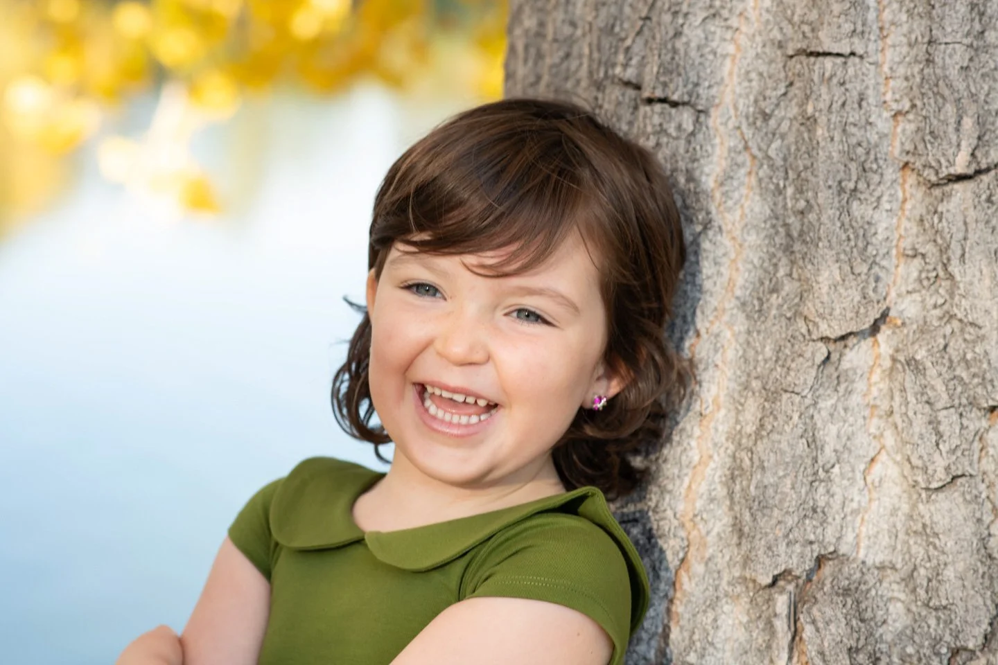 Young girl in a green dress laughing joyfully while leaning against a tree, capturing a natural and genuine smile for a family portrait by Kellie Henriksen Photography.