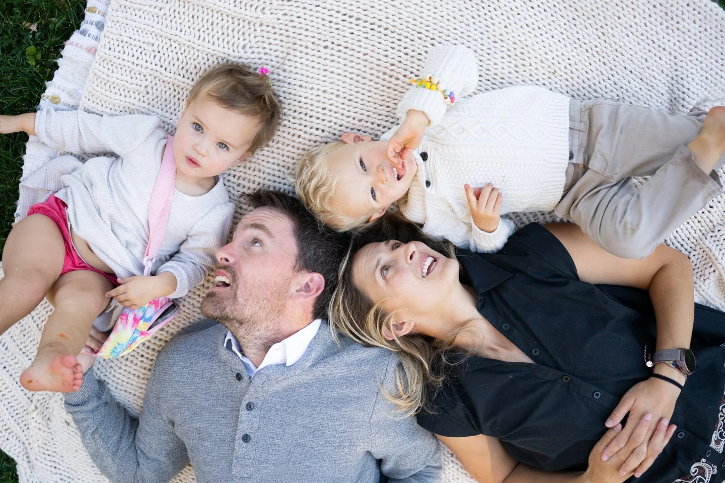 Mom, dad, 4 year old son and 2 year old daughter laying on a blanket on the lawn  smiling in a family portrait session by photographer Kellie Henriksen