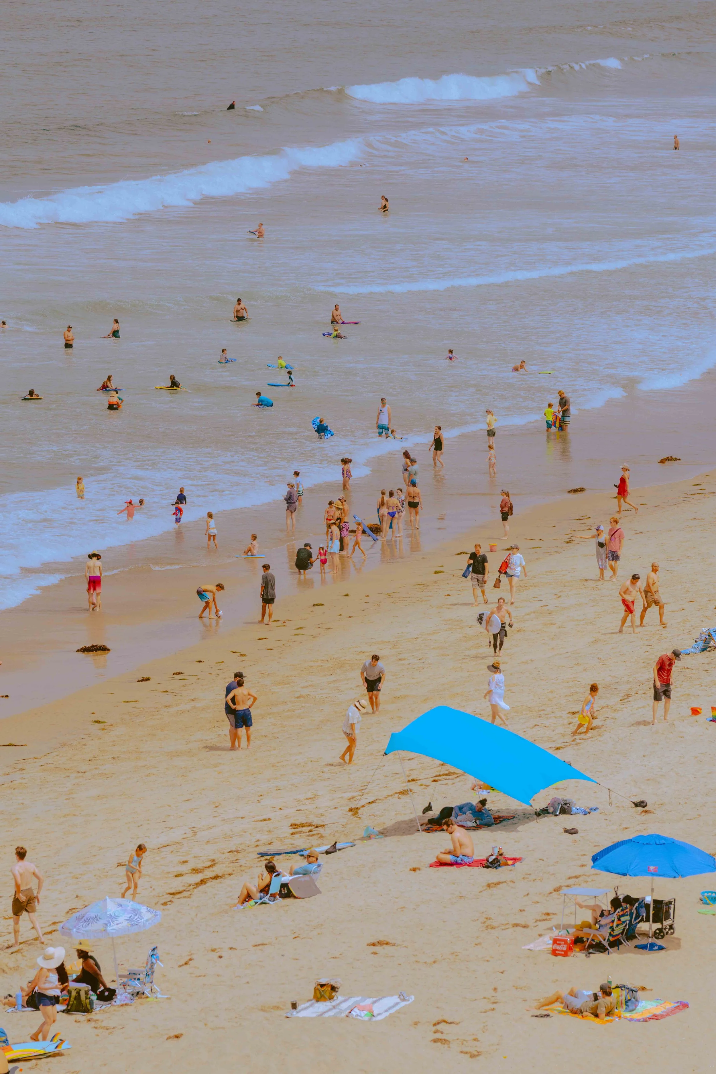 Beach scene with people swimming, sunbathing, and walking on the sand under umbrellas, with some in the water and others on the beach.