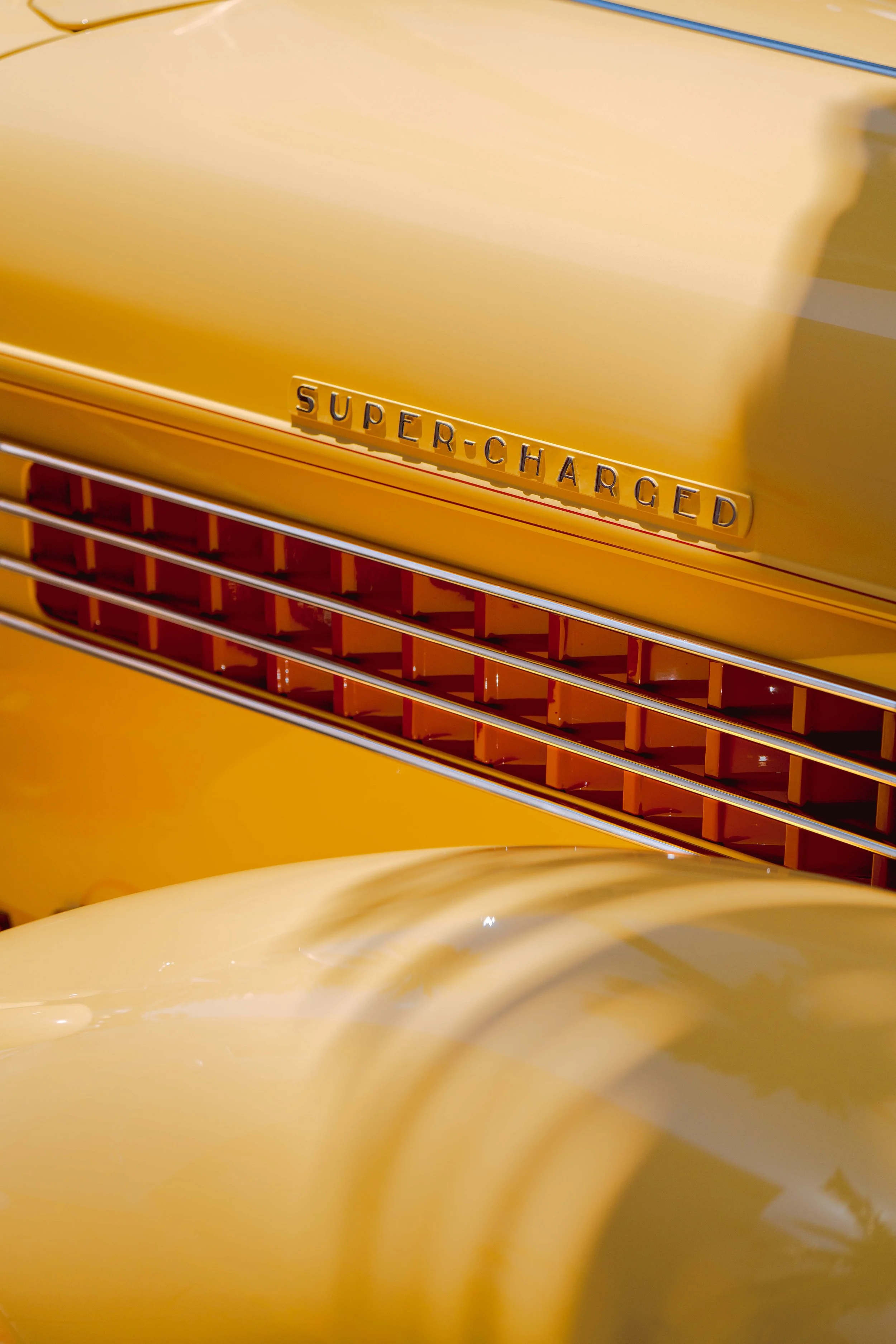 Close-up of the front grille of a yellow vintage car, with a badge that reads "Super-Charged."