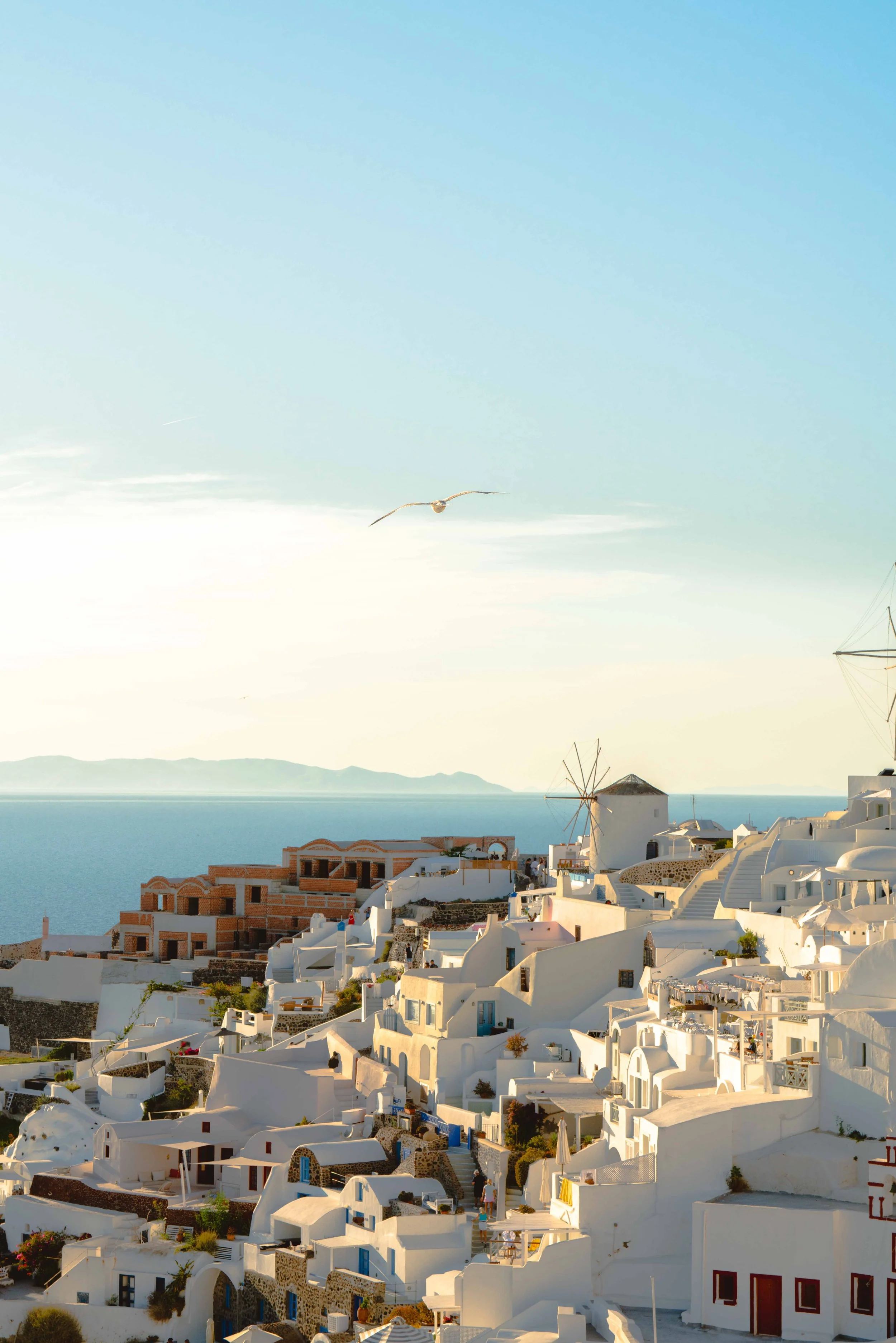 White-washed buildings with blue accents on a hillside overlooking the sea, with windmills and a seagull flying in the sky.