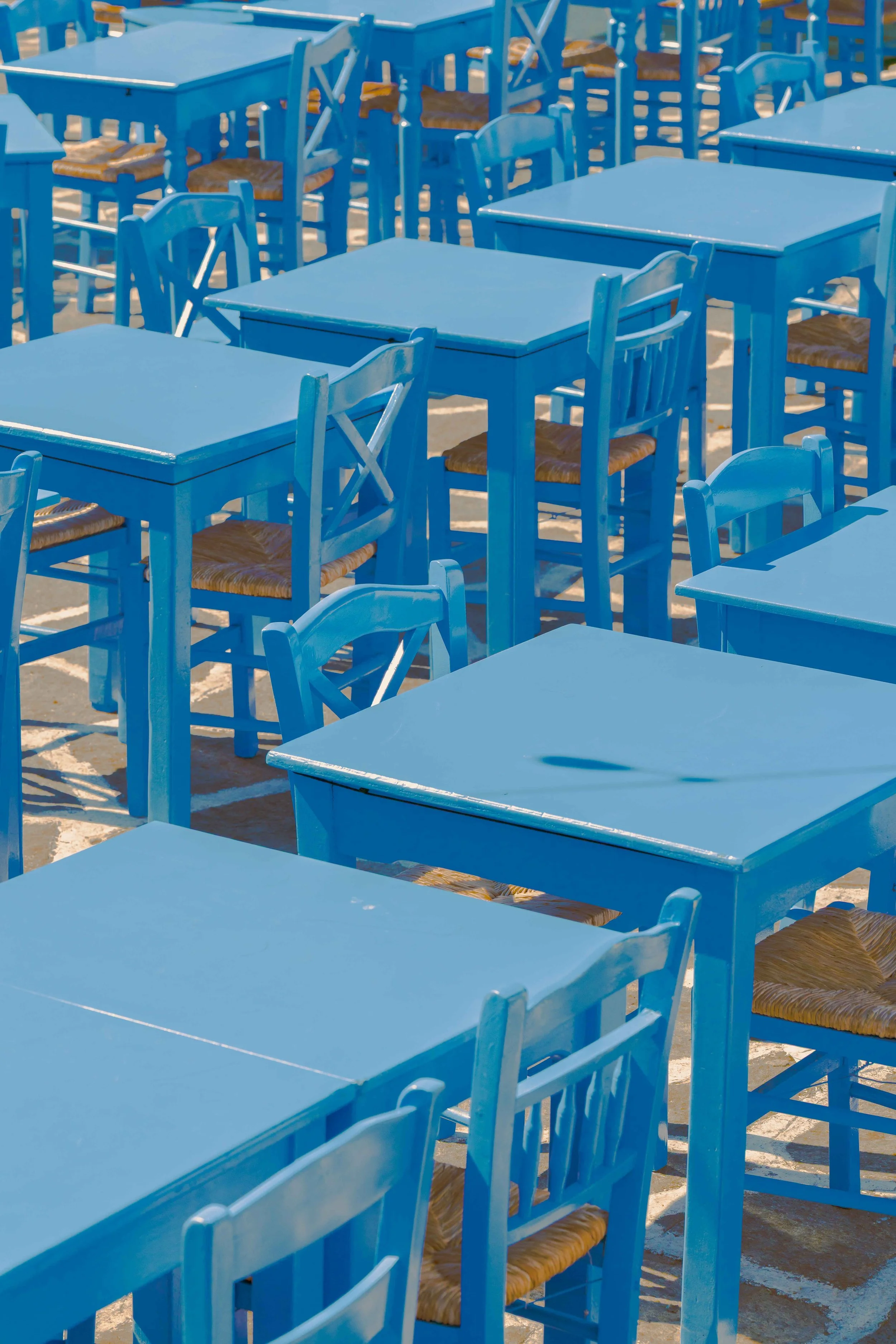 Multiple blue wooden tables and chairs with woven seats arranged in an outdoor setting, likely on a beach or patio.