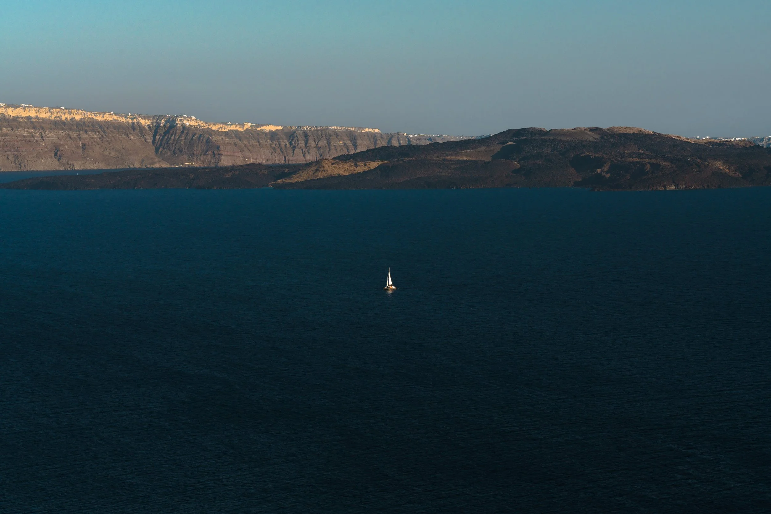 A sailboat floating in a calm, dark blue sea with rugged, dark brown and green islands and distant cliffs in the background under a clear sky.