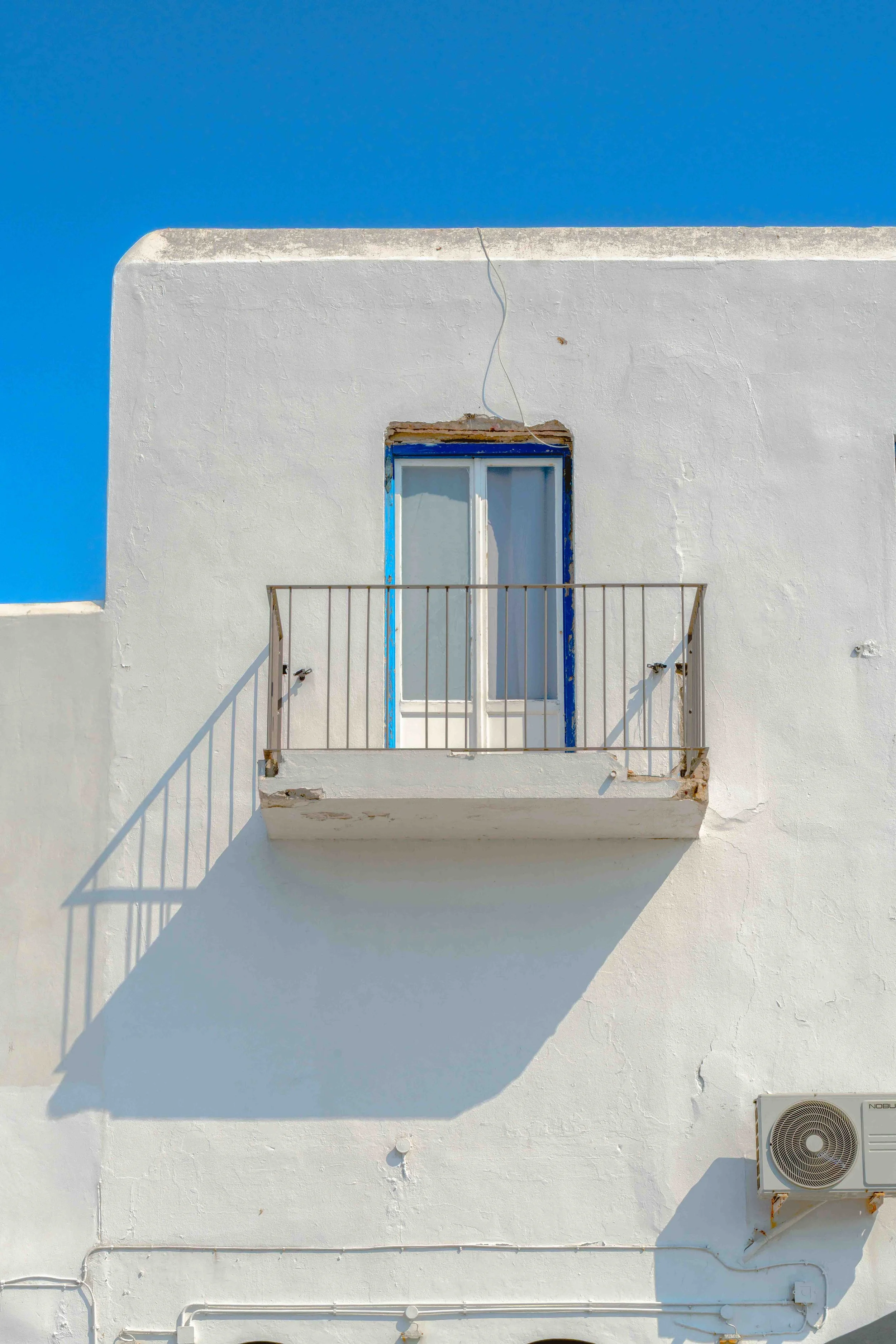 White building exterior with a small balcony, a closed white door with blue trim, above which is a small piece of roofing, and a shadow cast below the balcony. An air conditioning unit is mounted on the wall.