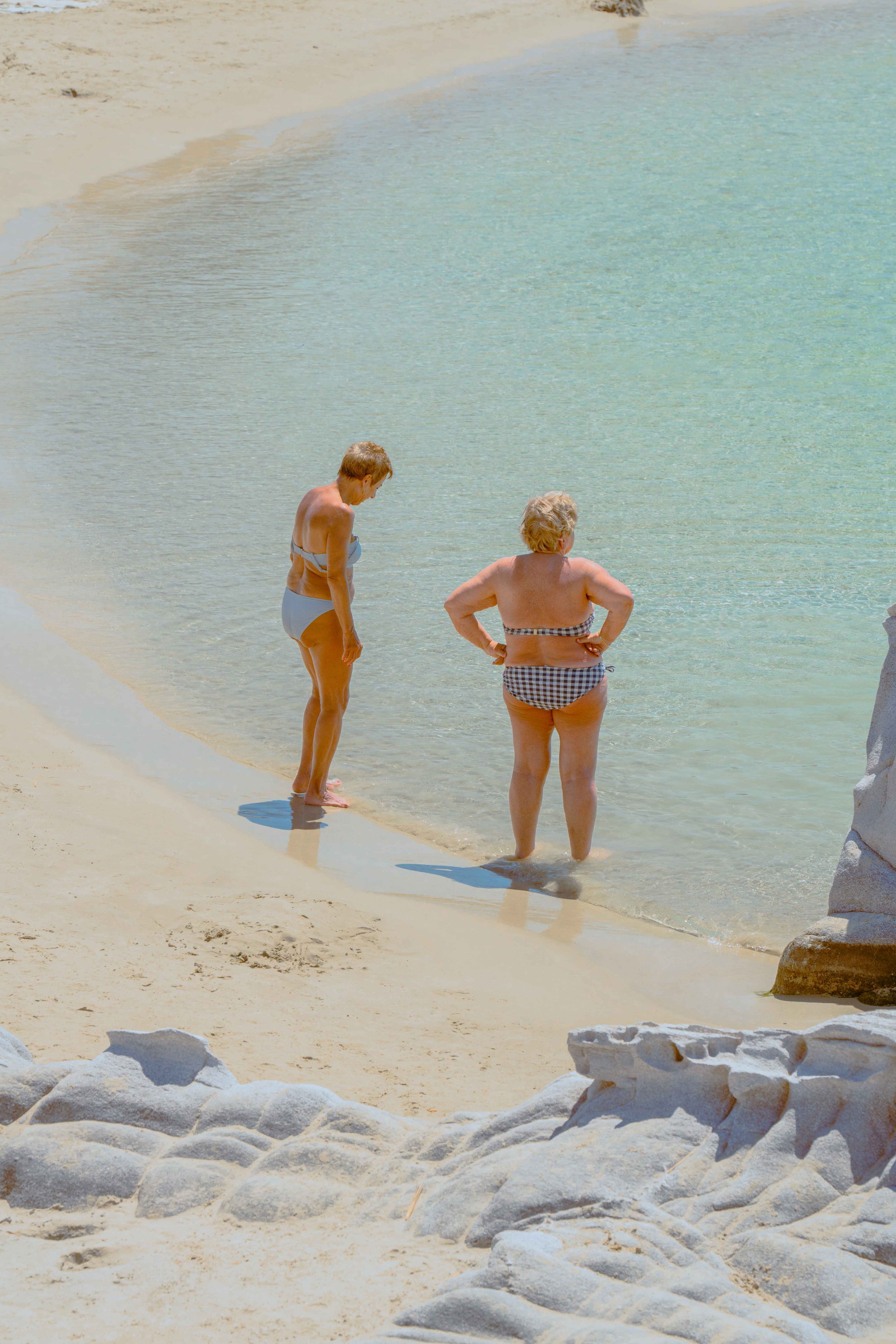 Two elderly women in swimsuits standing by the shoreline of a beach, with one woman looking down into the water and the other with hands on hips facing the sea. The sand is light-colored with some rocks in the foreground, and the water is calm and cl