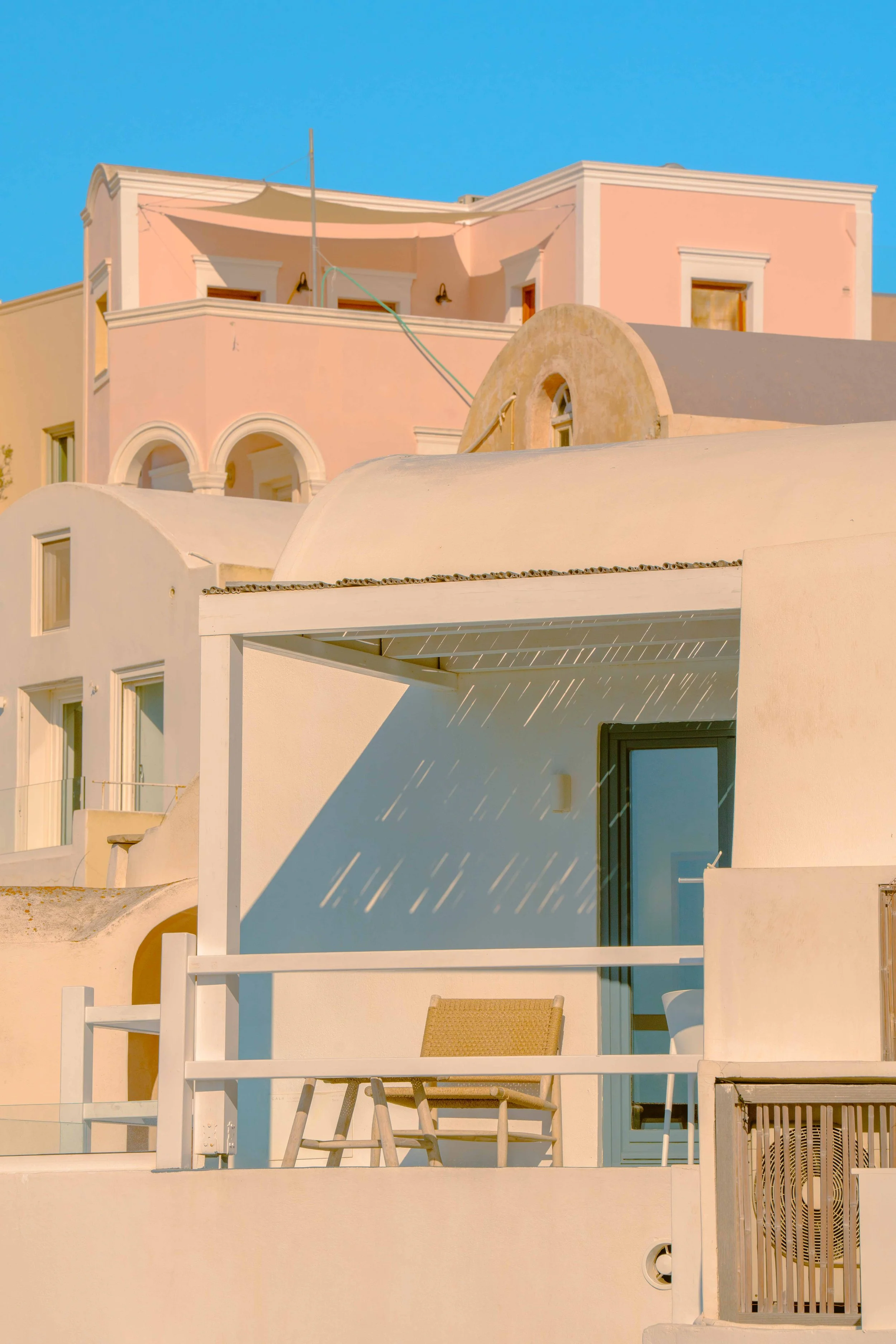 A building with a pastel pink and white facade, featuring balconies, arched windows, and a terrace under shade, set against a bright blue sky.