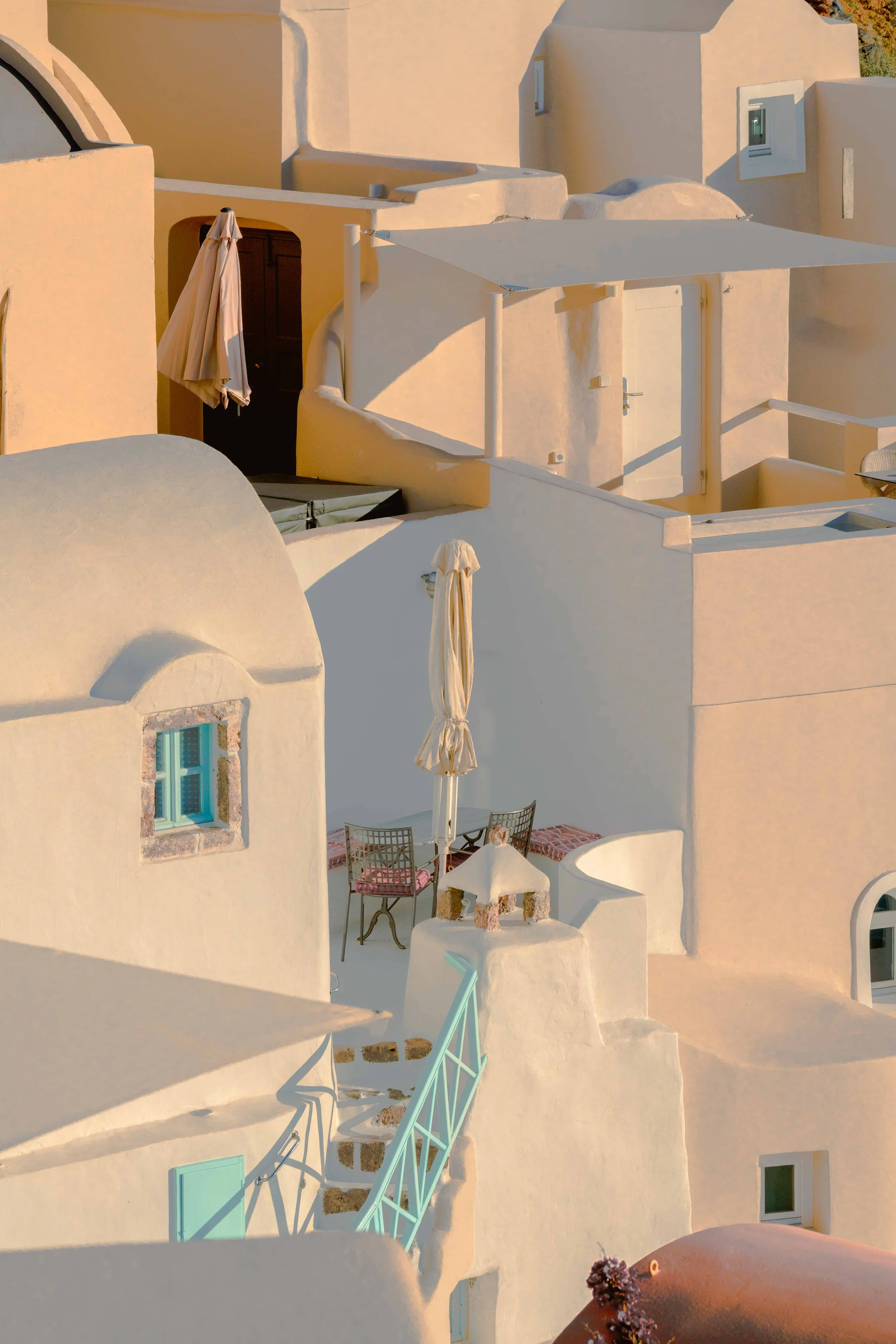 A white-washed building with rounded edges, a small window with turquoise shutters, outdoor seating, an umbrella, and stairs with a railing in a Mediterranean setting.