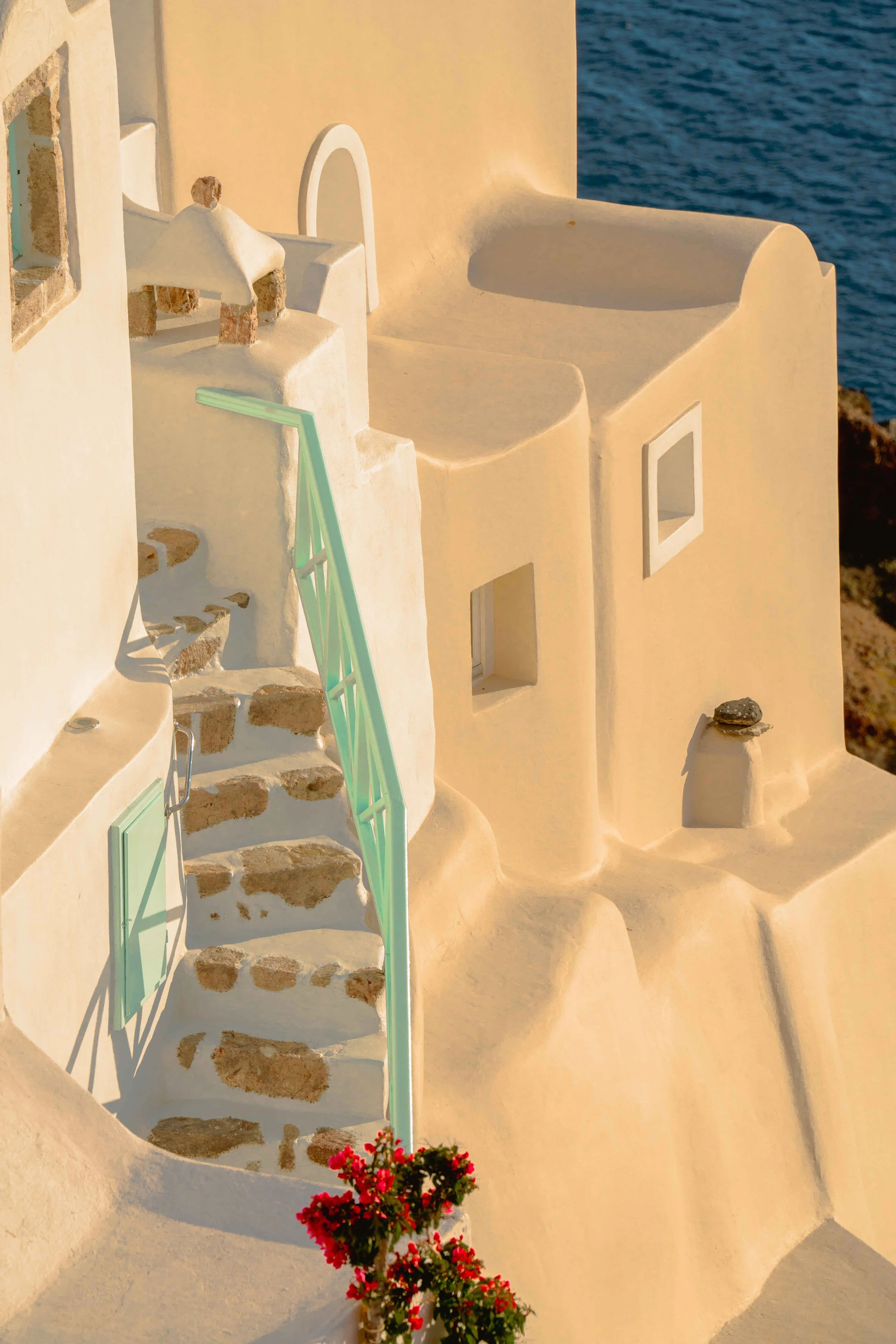 White buildings with rounded edges and small windows, staircase with green railing, and potted pink flowers, overlooking the sea.