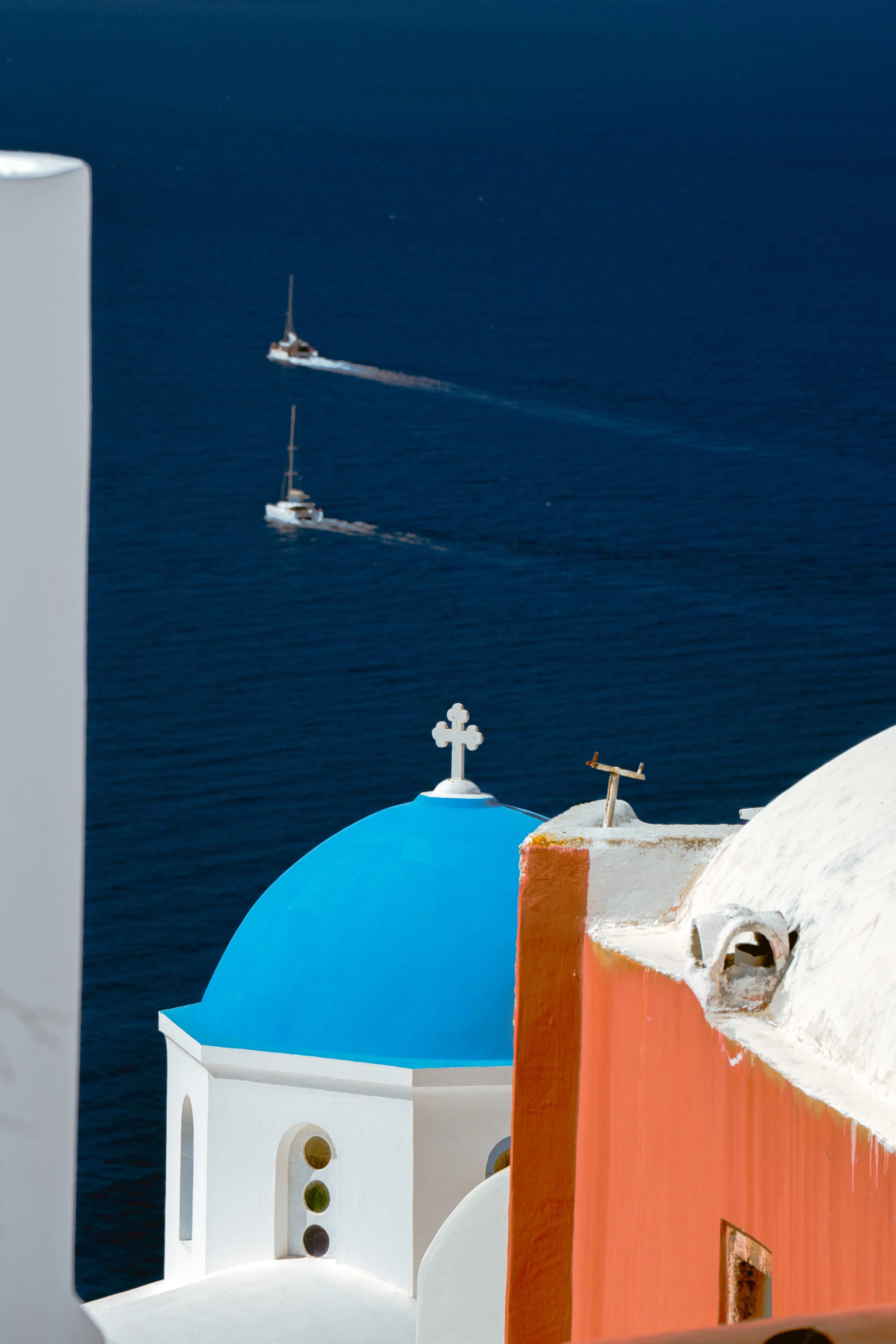 White building with blue dome roof, orange wall, and white cross ornament against the blue sea background in Greece.