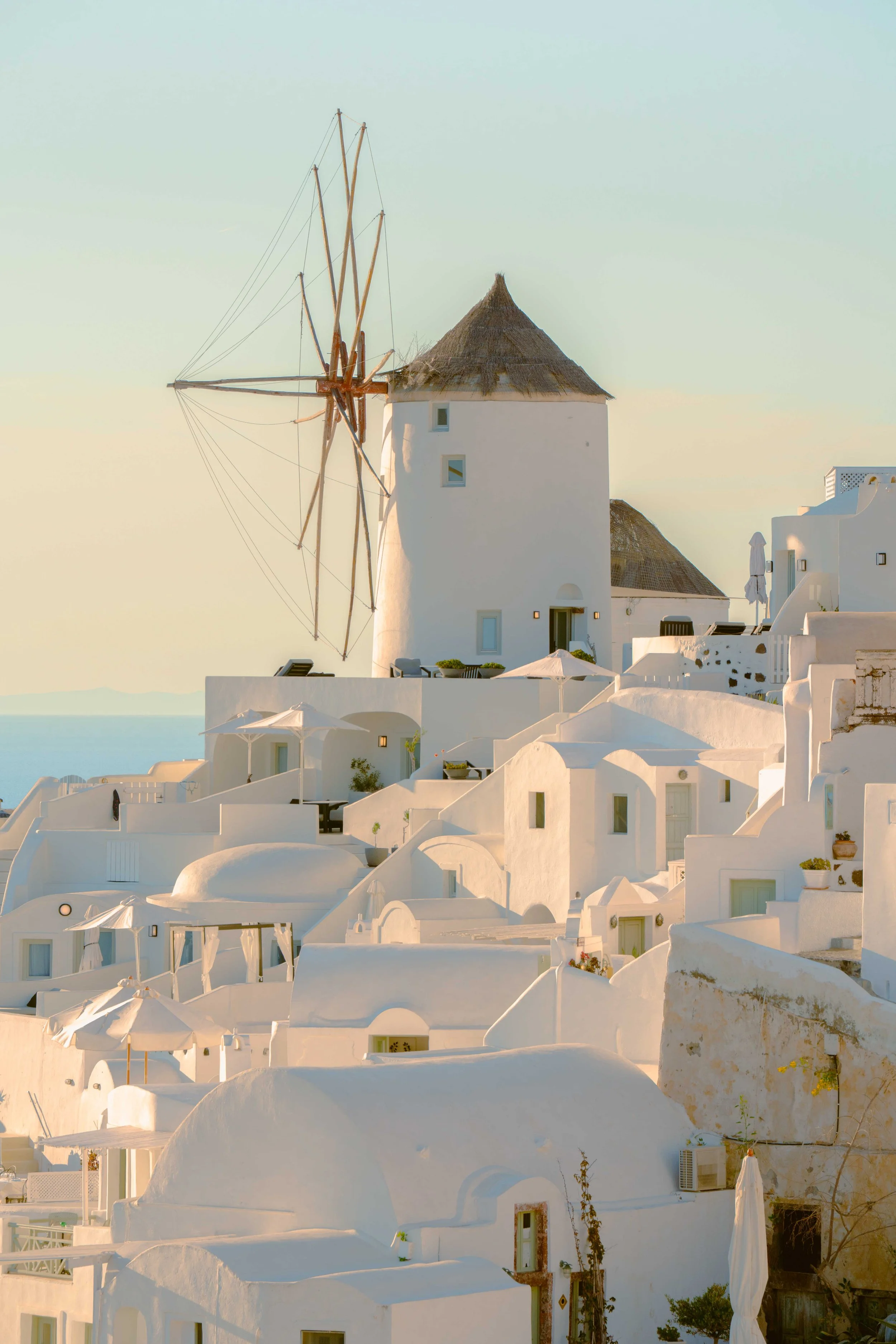 A picturesque view of white buildings with rounded edges and flat roofs typical of Santorini, Greece, under a pale sky. A traditional windmill with wooden blades and a thatched roof stands prominently on a hill among the structures.