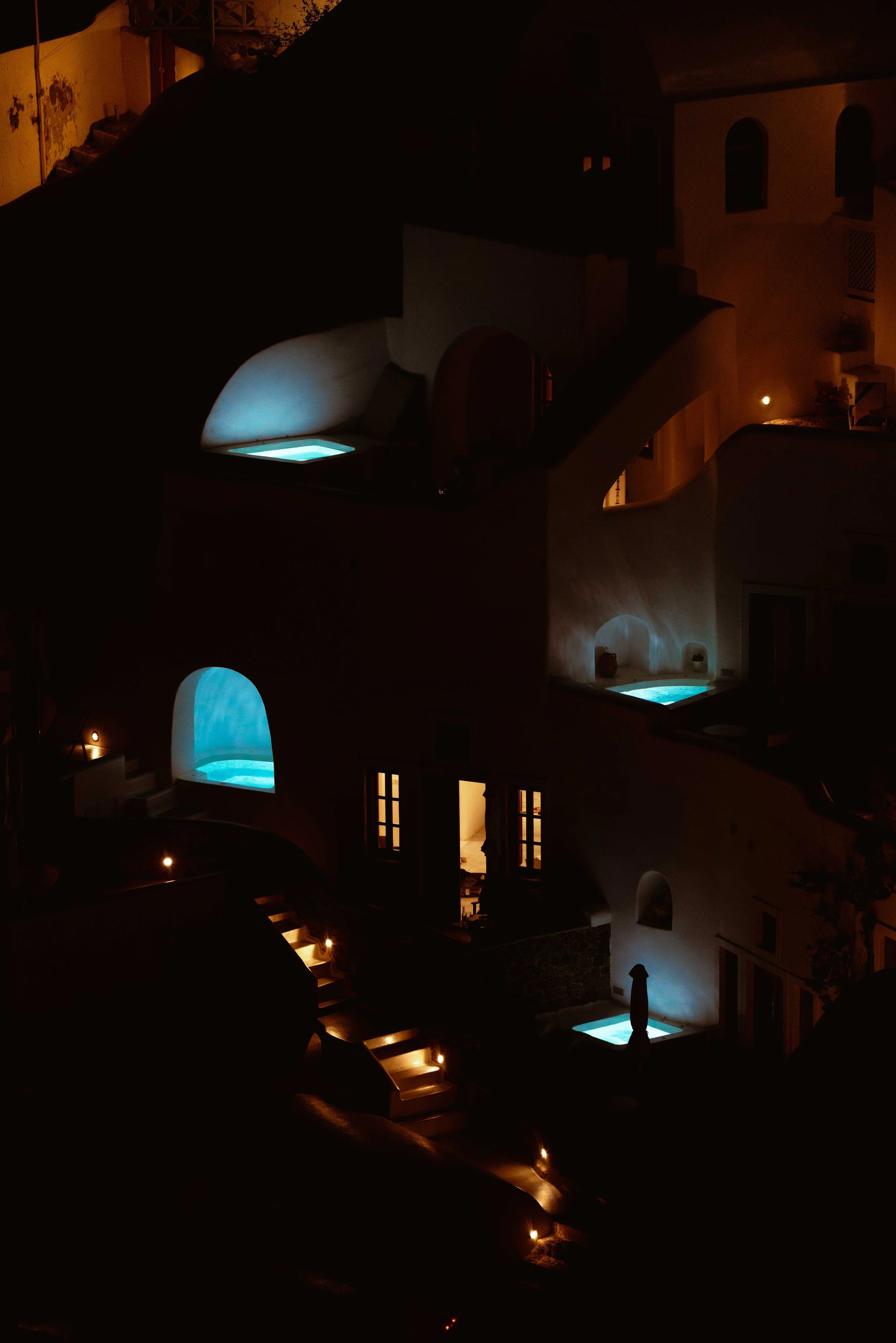 Nighttime view of a Mediterranean-style house with illuminated swimming pools and outdoor stairs, featuring white walls and arched doorways.