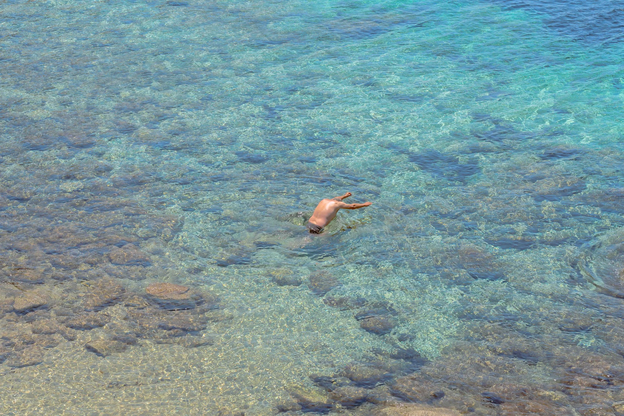 A person swimming in clear blue water near a rocky beach shoreline.