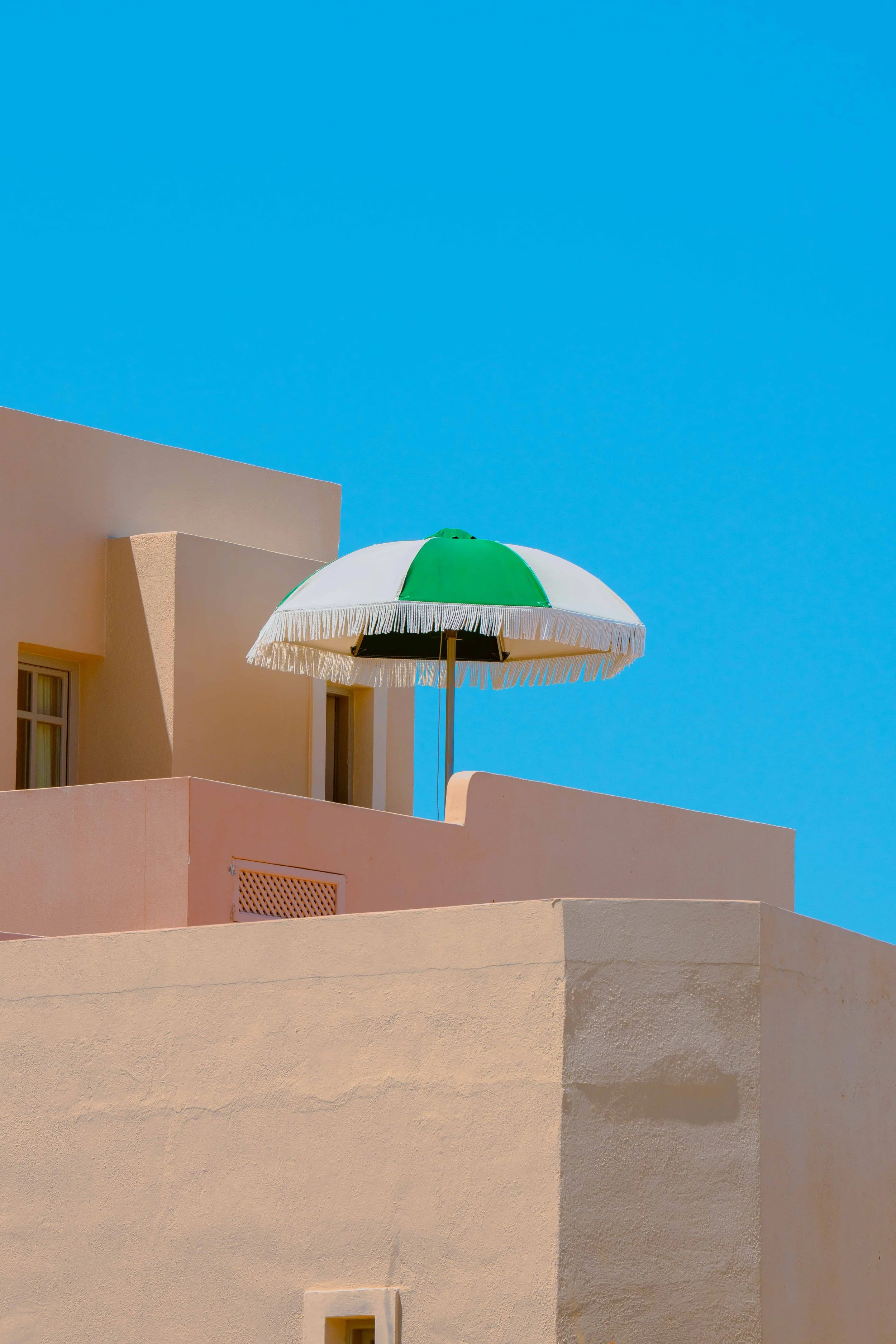 A pastel pink building with minimalistic design, featuring small windows and a beige air vent, topped with a white and green beach umbrella against a clear blue sky.
