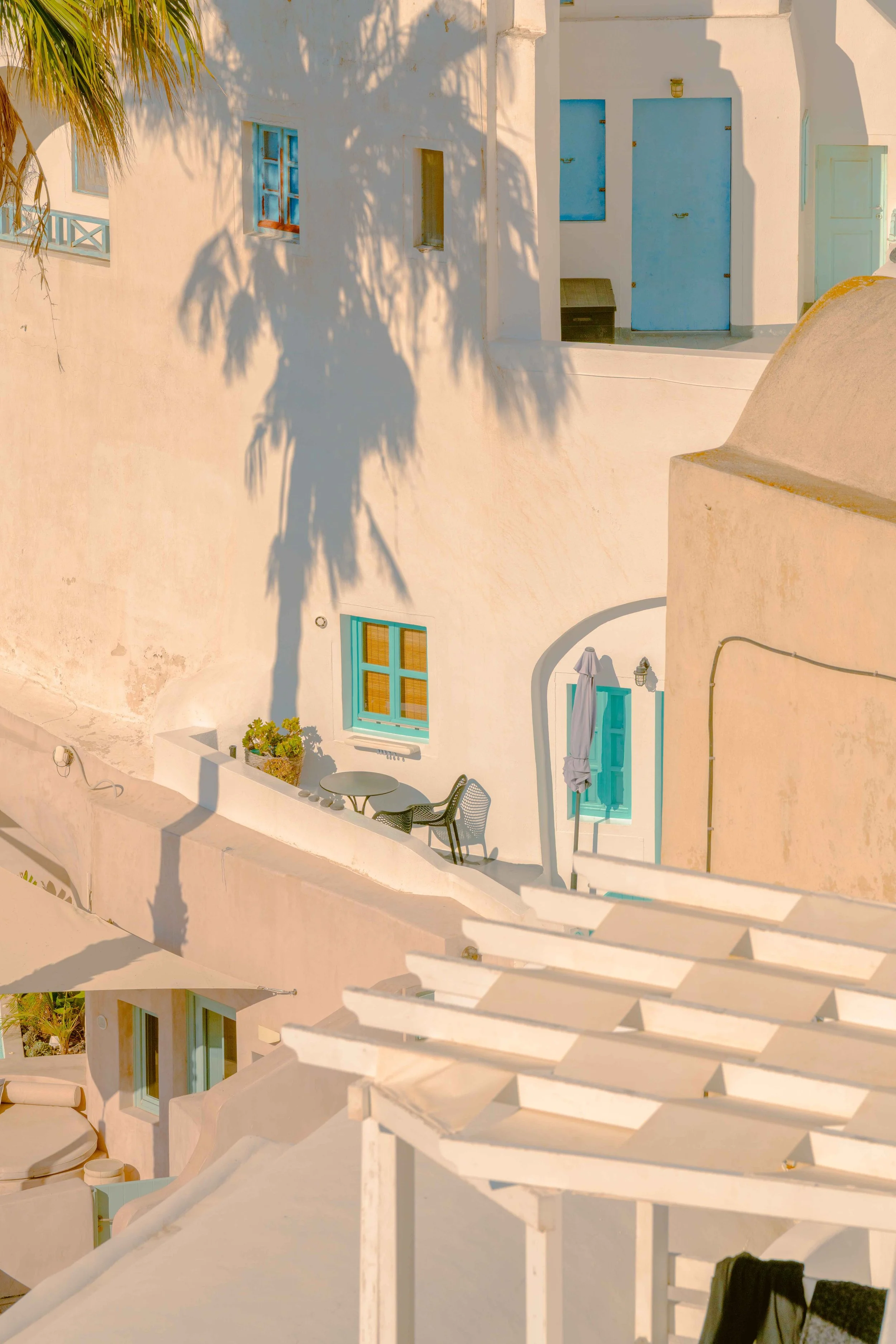 White Mediterranean-style building with blue doors and windows, shaded by palm tree, featuring outdoor seating and decorative elements.
