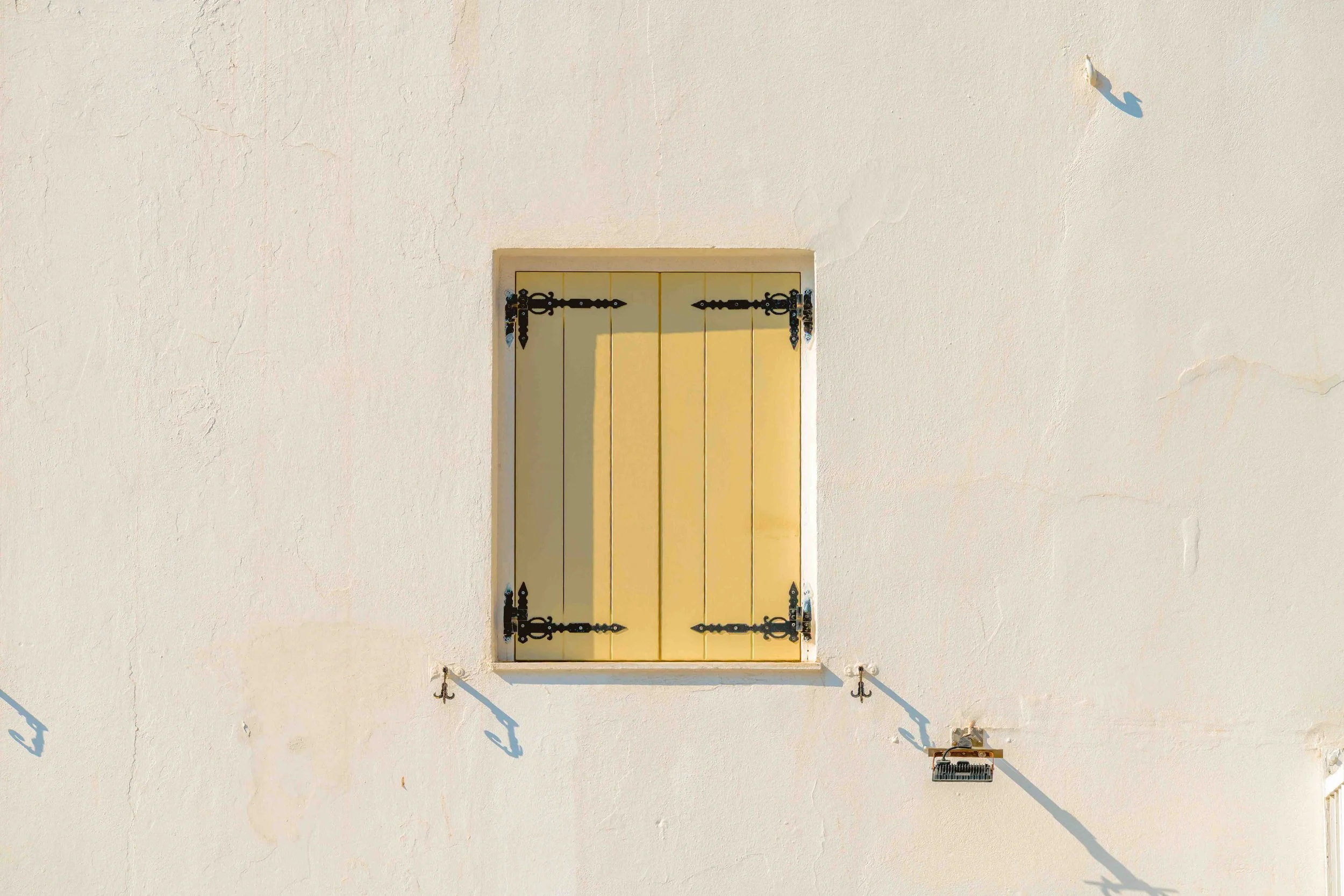 A closed yellow window with decorative black iron hinges on a white stucco wall, with shadows cast by the hinges and window on the wall.