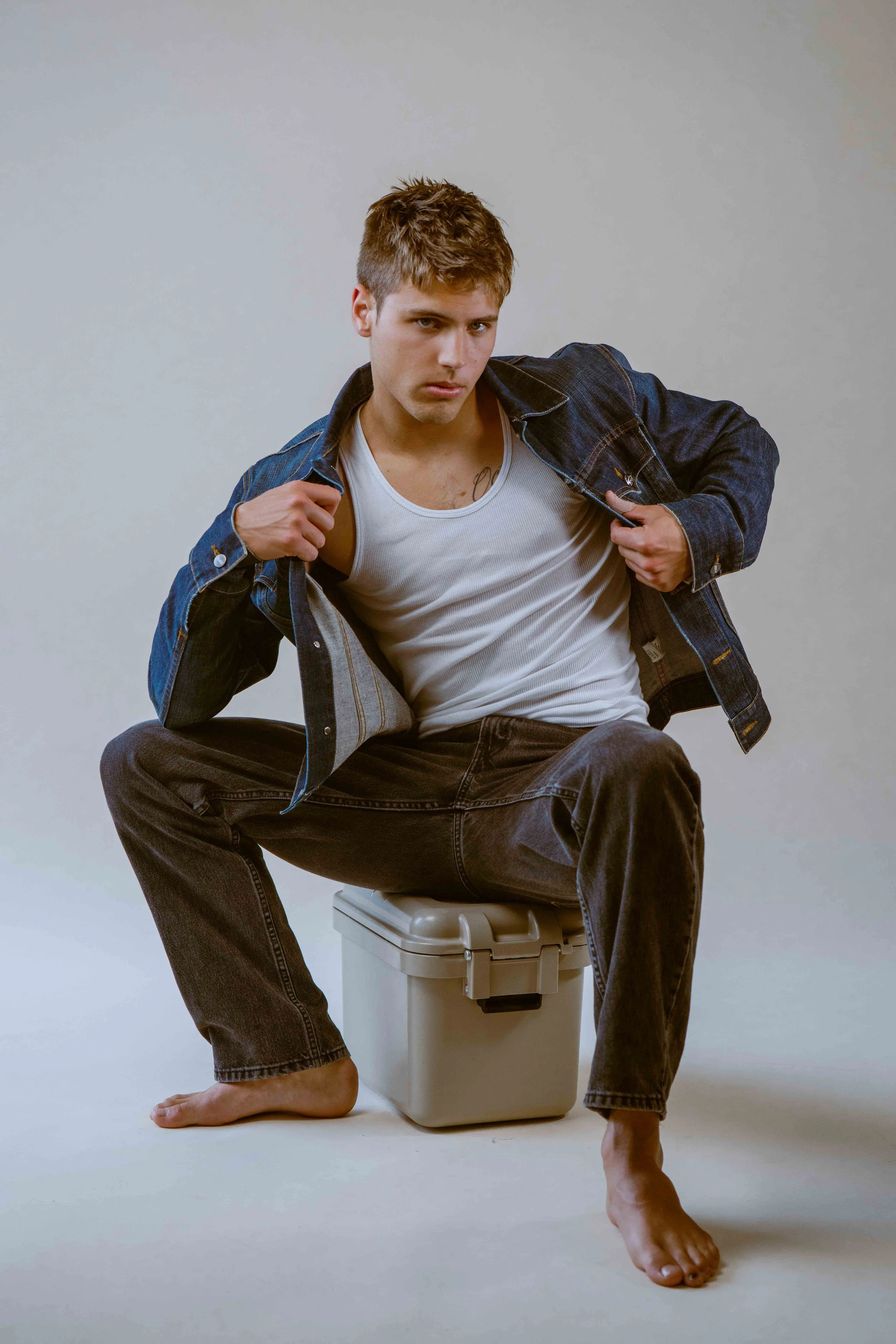 A young man with short hair and light skin, wearing a white tank top and a denim jacket, sitting on a white plastic storage container. He is offset to the left, resting his right arm on his chest and adjusting his jacket with his left hand. His right foot is barefoot and resting on the floor, while his left foot is also bare and placed on the floor. The background is a plain, light-colored wall.