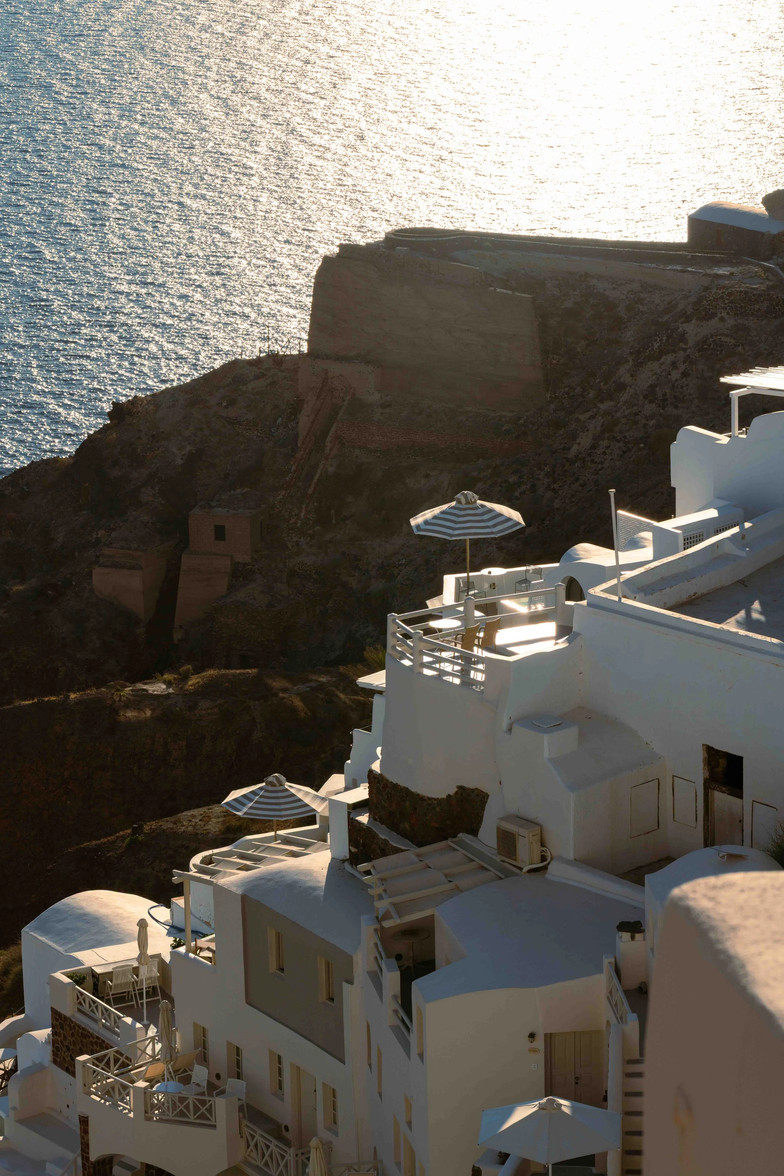 White buildings with terraces and striped umbrellas on a hillside overlooking the sea in Santorini, Greece, during sunset.