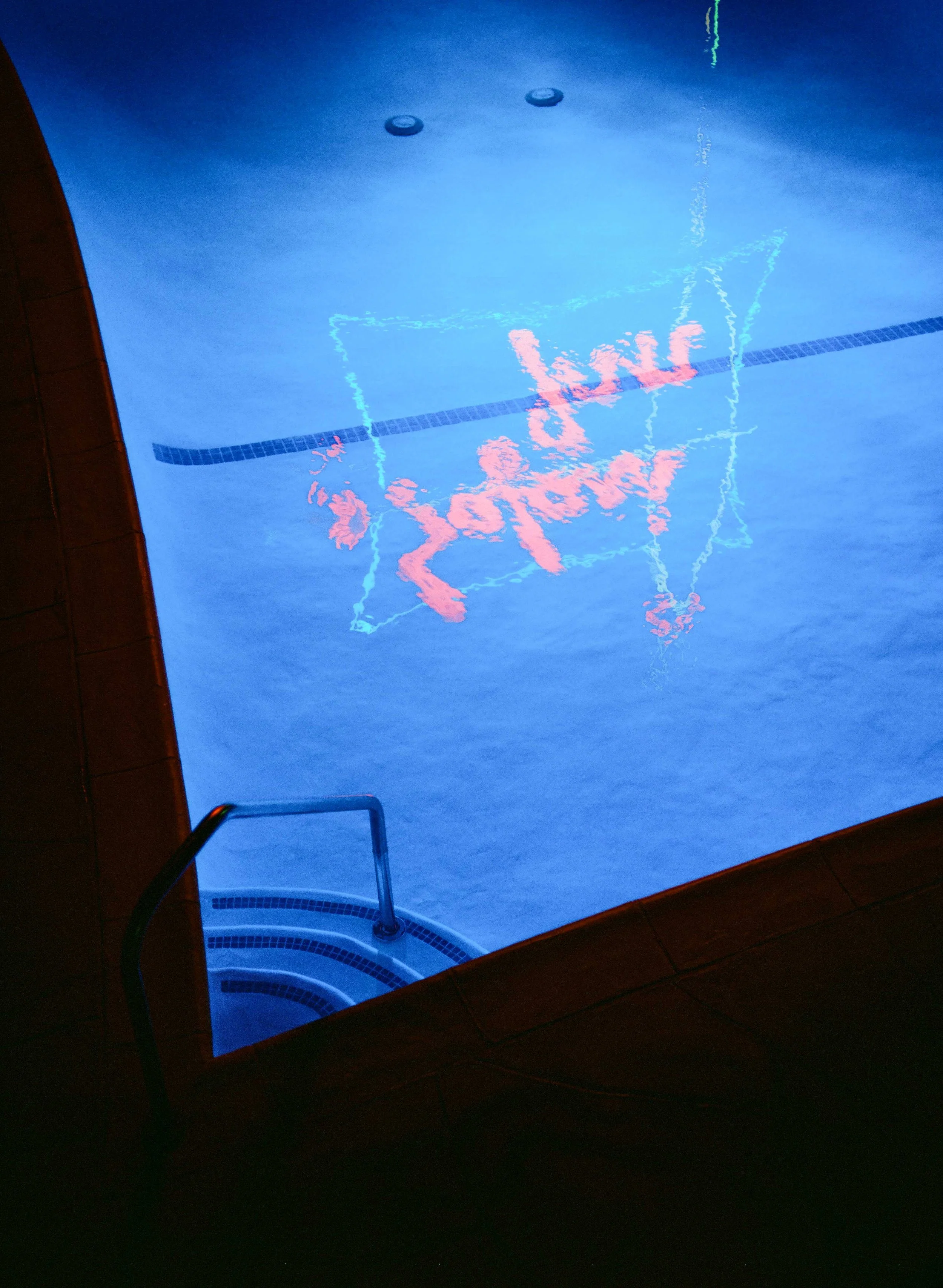 Indoor swimming pool with blue water, staircase, and reflection of a neon sign on the water's surface.
