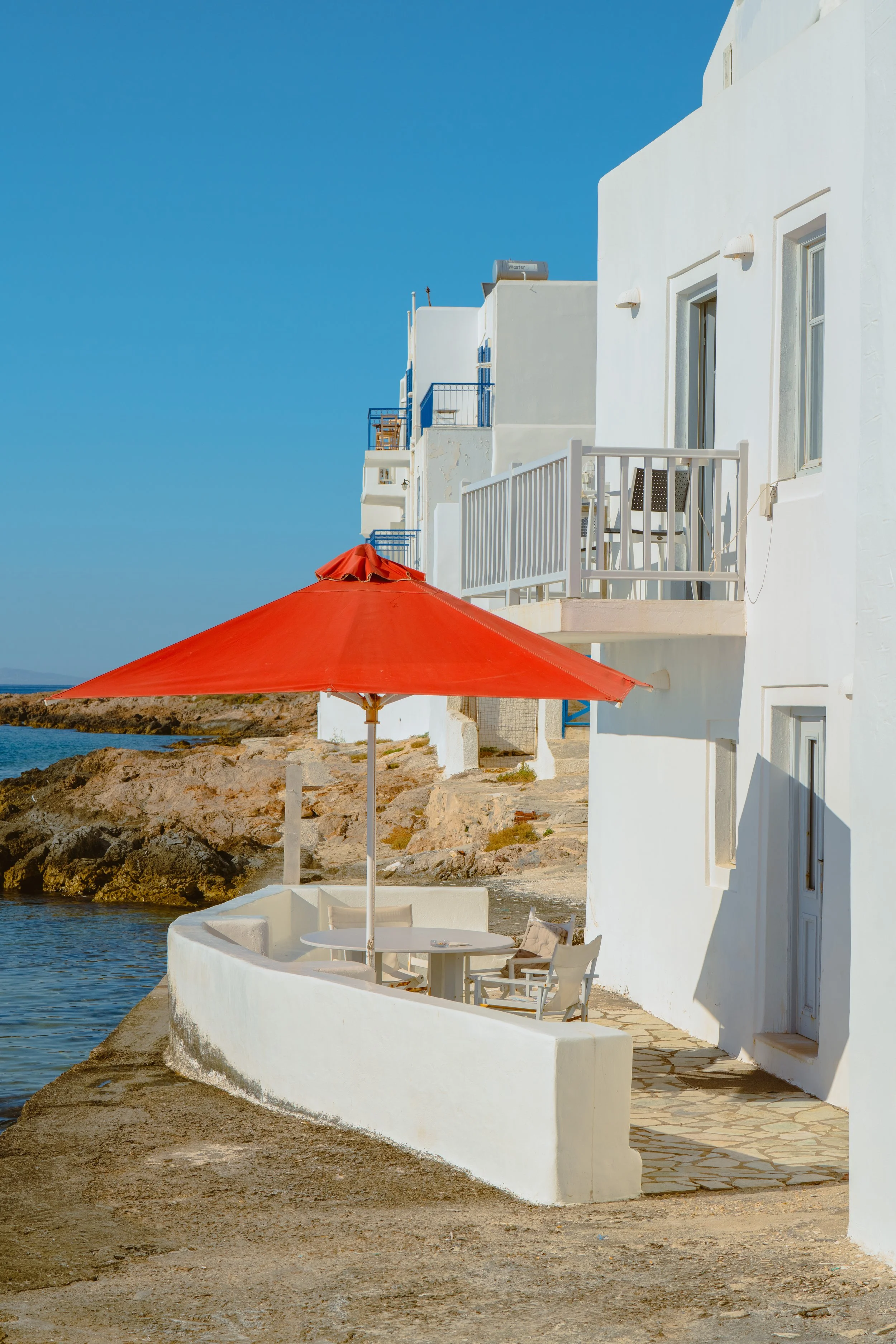 White buildings with balconies and windows, a red umbrella shading a patio with chairs, rocky shoreline, and a clear blue sky.
