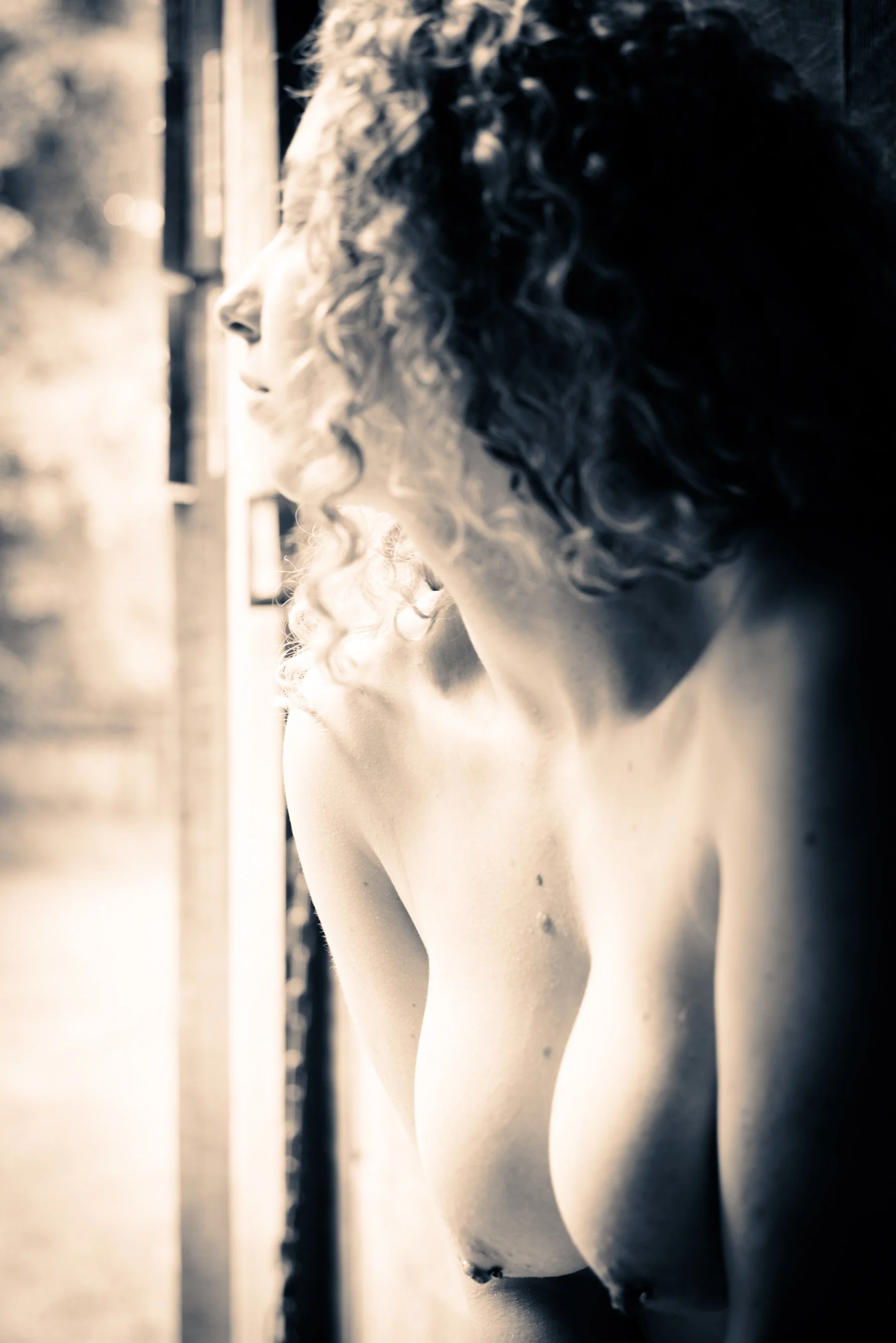 A black-and-white photo of a woman with curly hair gazing out of a window, with sunlight illuminating her face and shoulder.