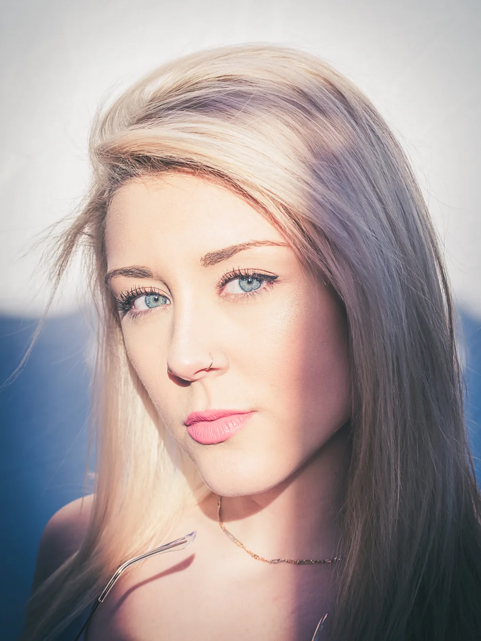 Close-up portrait of a woman with blonde and purple hair, blue eyes, and a nose ring, outdoors in sunlight.