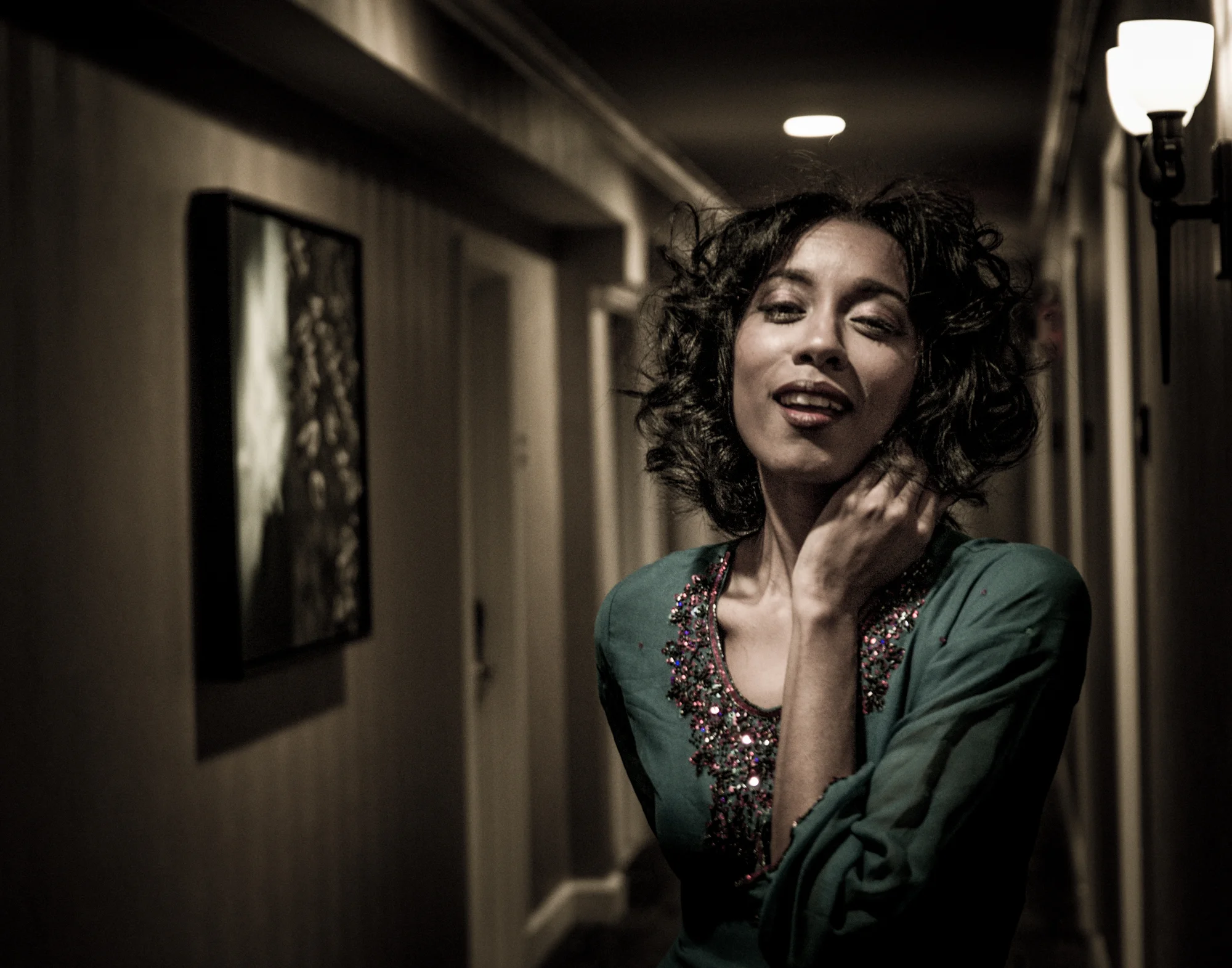 A woman with curly dark hair posing in a dimly lit hallway, wearing a green blouse with colorful embroidery around the neckline.