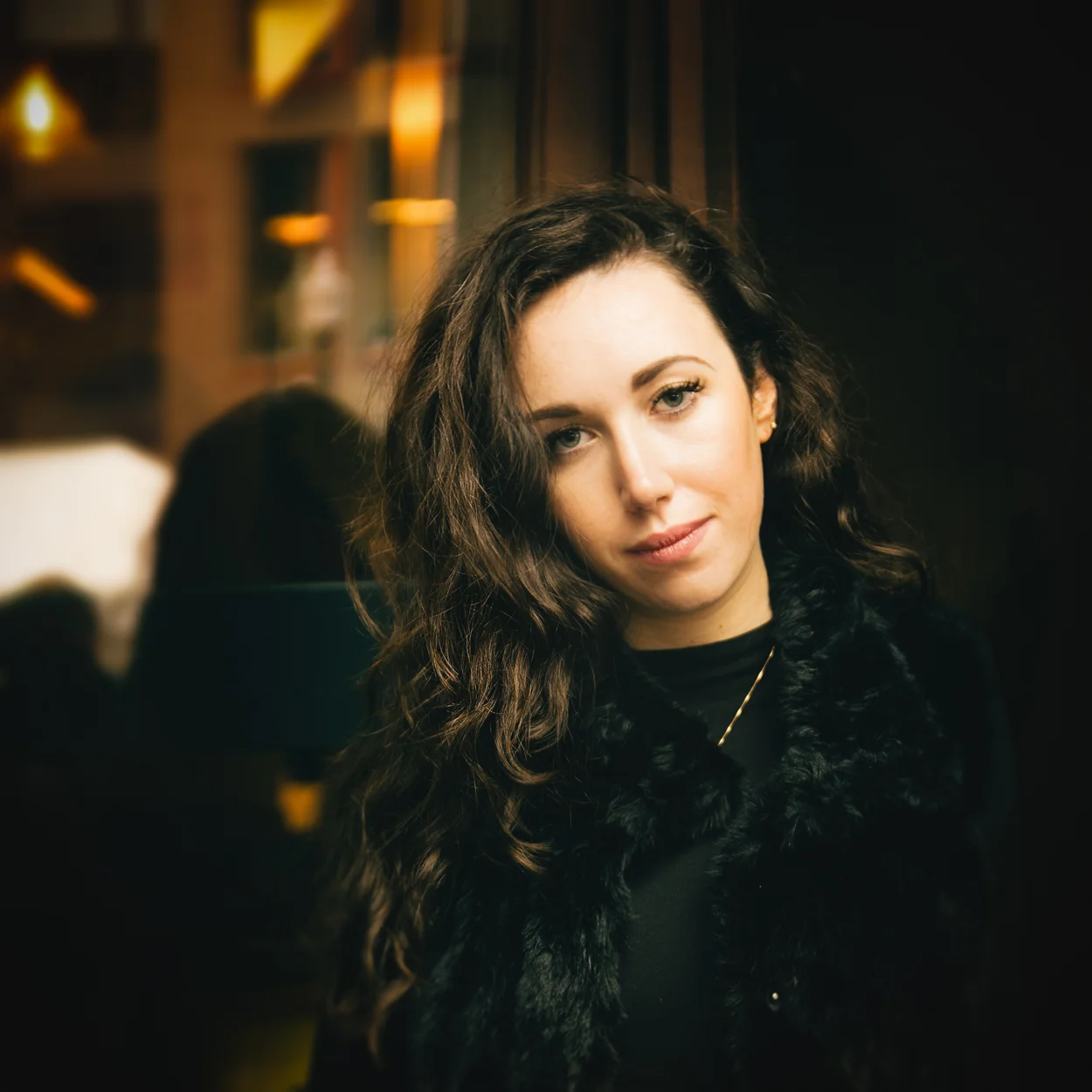 A young woman with long, dark, curly hair posing indoors with warm lighting, wearing a black top and a black fur vest.