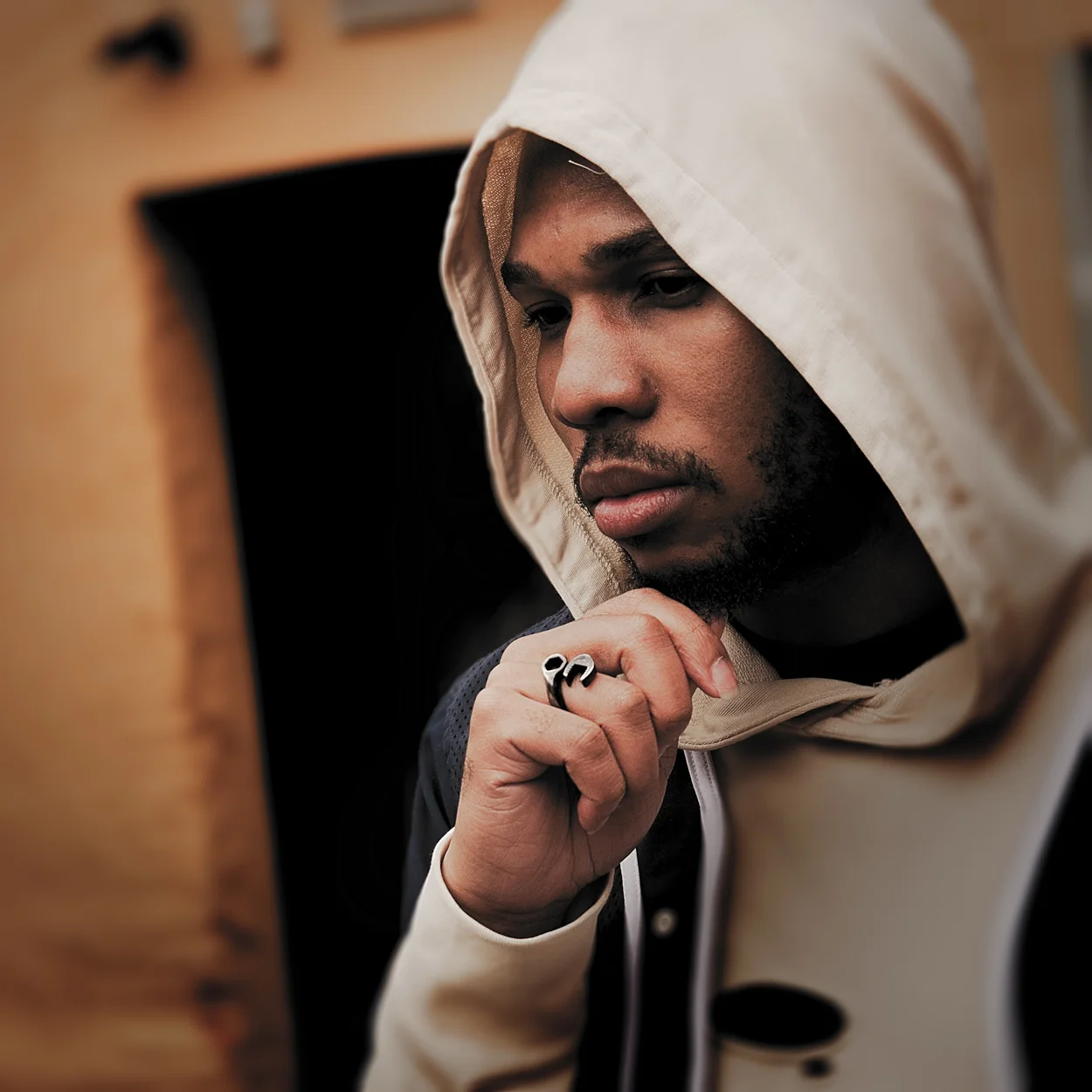 A young man wearing a beige hoodie and a black jacket, with his hand near his chin, looking thoughtfully at a computer or mirror.