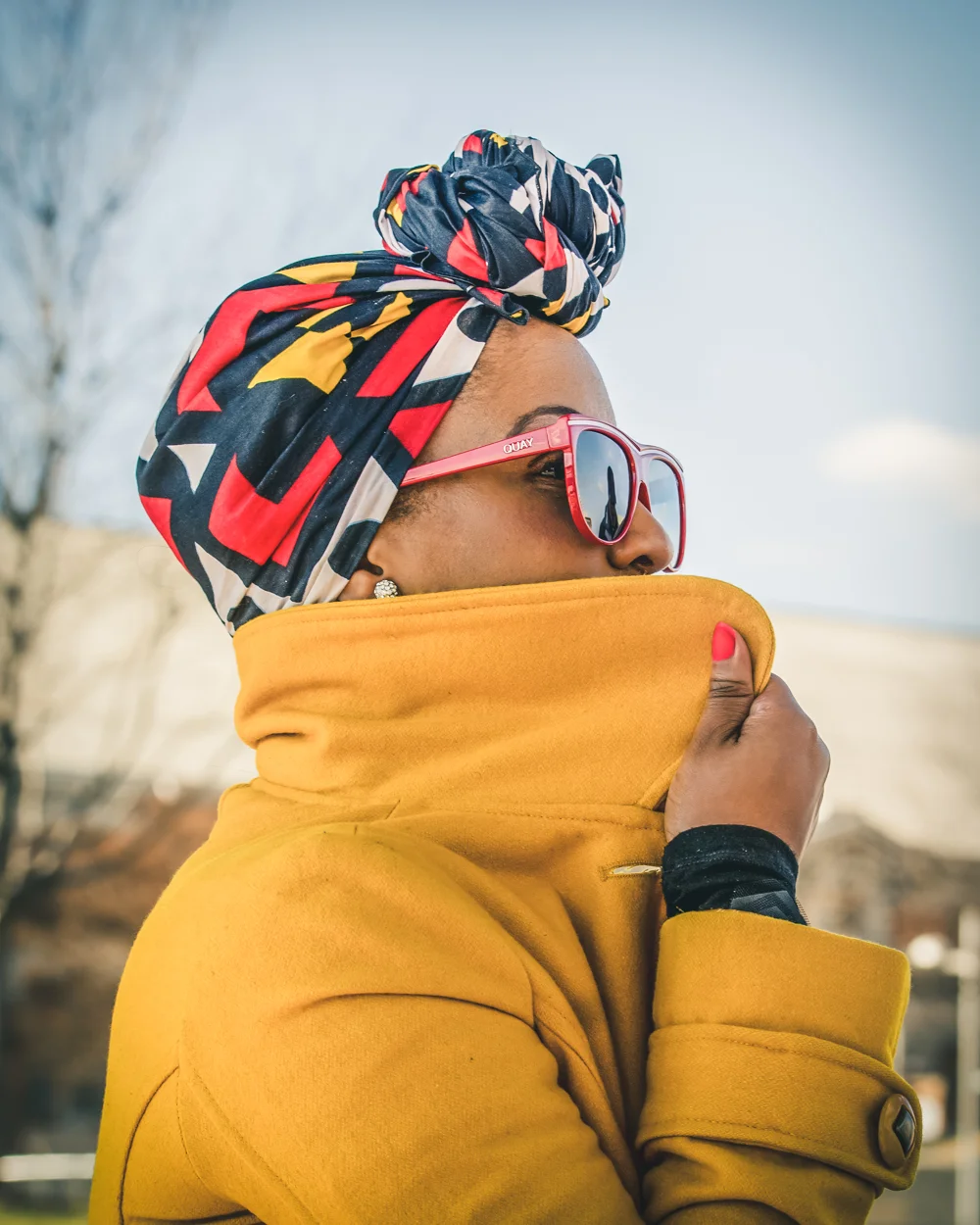 A woman wearing a colorful headwrap, red sunglasses, and a yellow coat, standing outdoors during daytime.