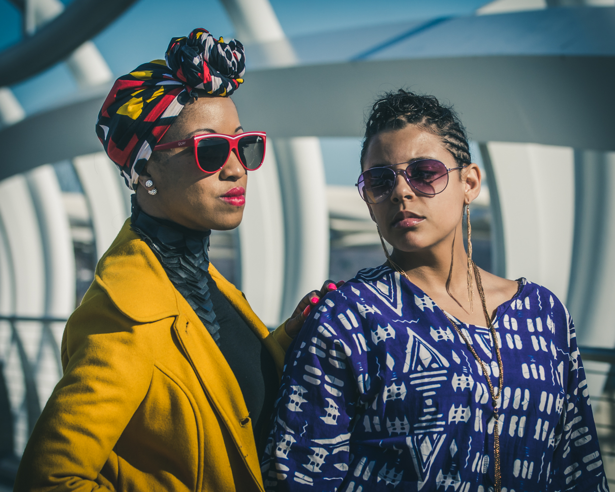 Two women wearing sunglasses and colorful clothing stand close together outdoors with modern architecture in the background.