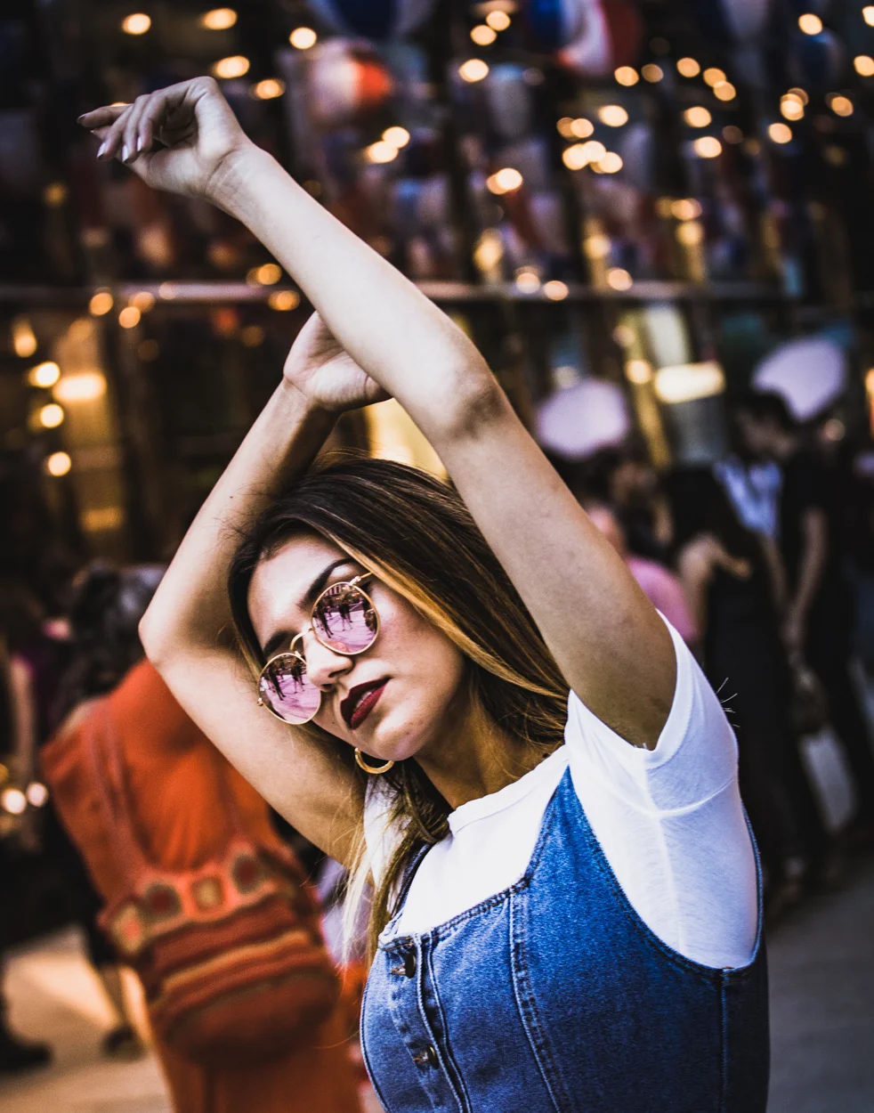 A young woman with long dark hair, wearing pink sunglasses, a white t-shirt, and a denim overall, is dancing with her arms raised in a lively indoor setting filled with people and warm lighting.