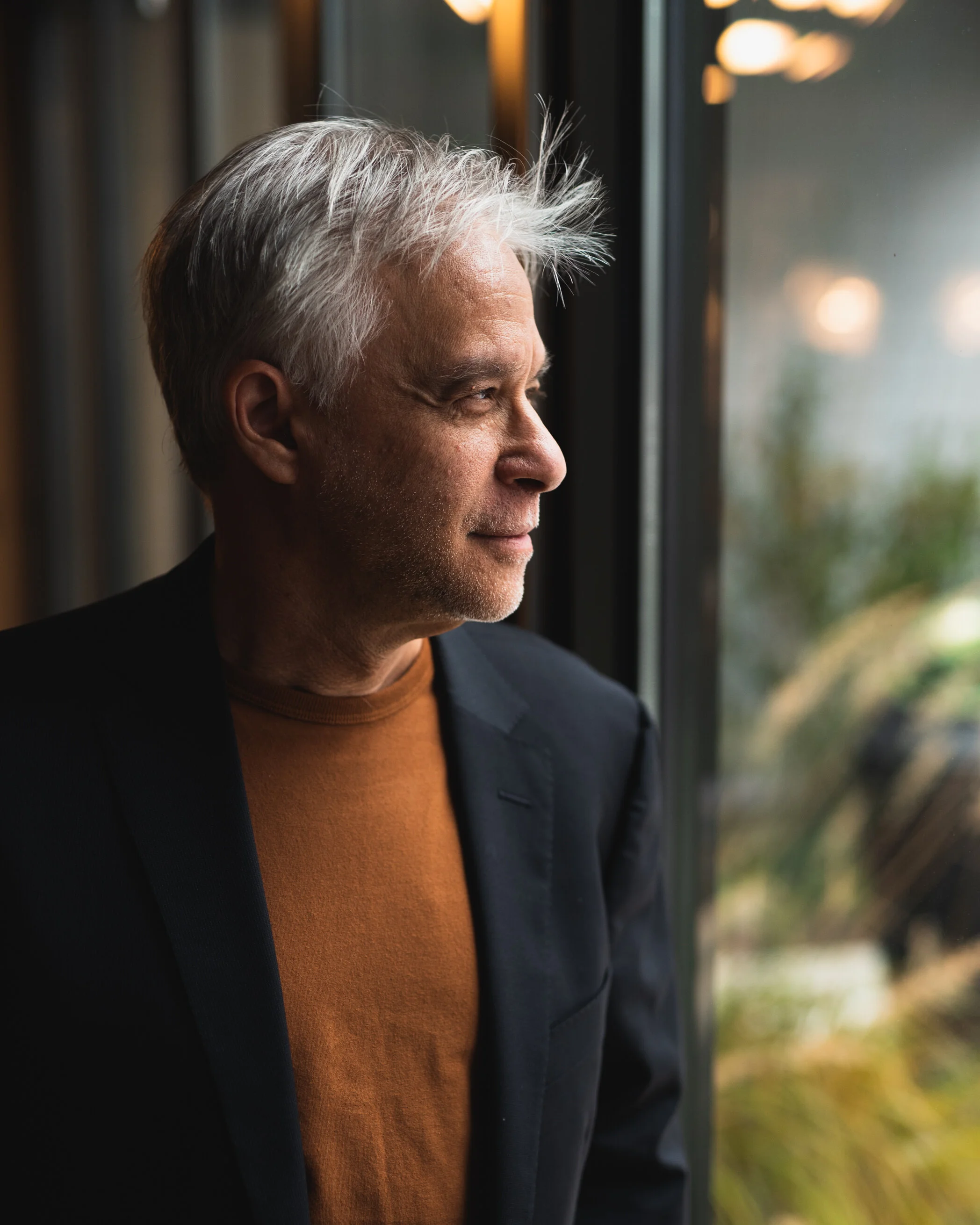 A middle-aged man with gray hair gazes thoughtfully out a window, dressed in a dark blazer and an orange shirt.