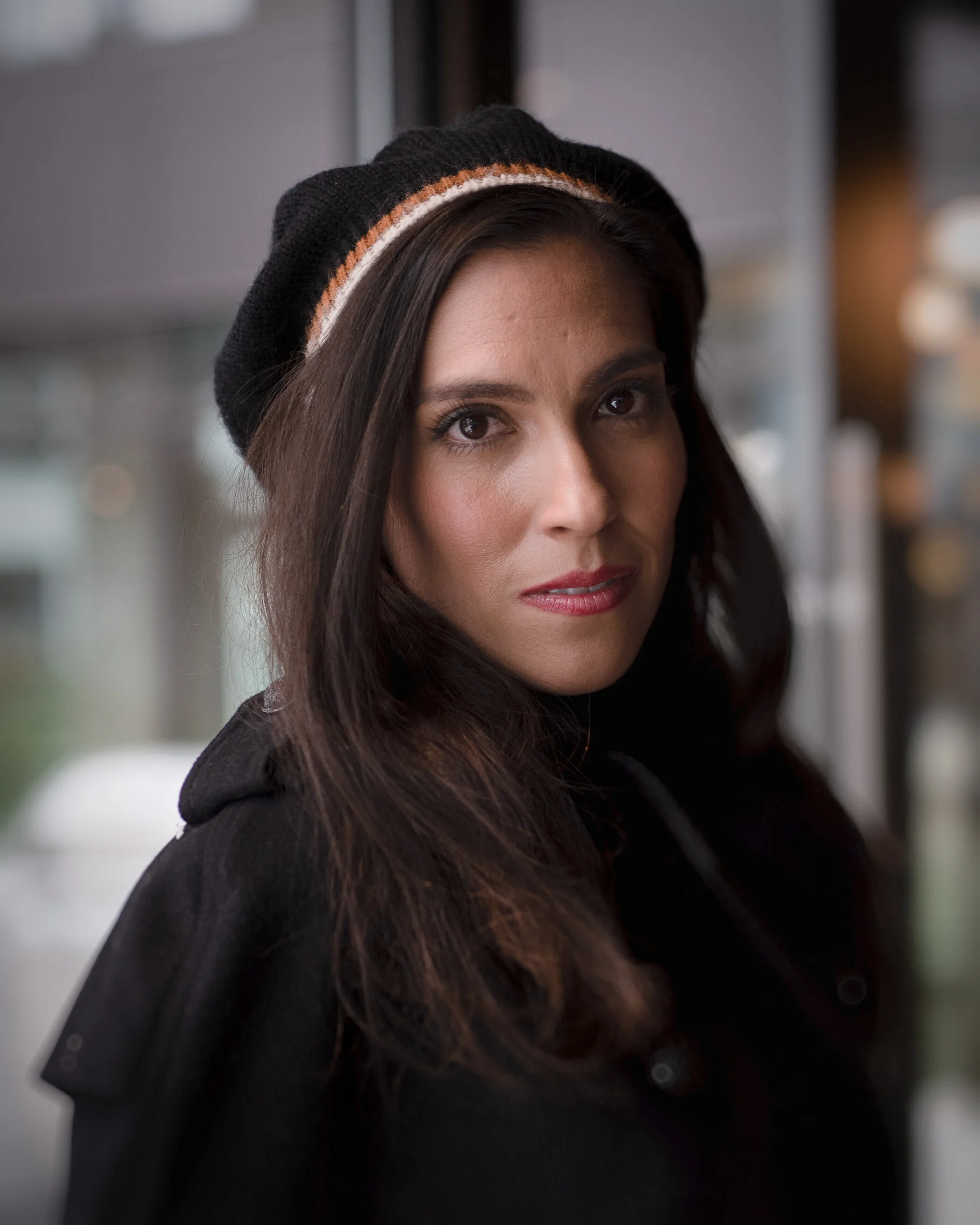 A woman with dark hair wearing a black beanie hat and a black coat, standing indoors near a window with a blurred background.