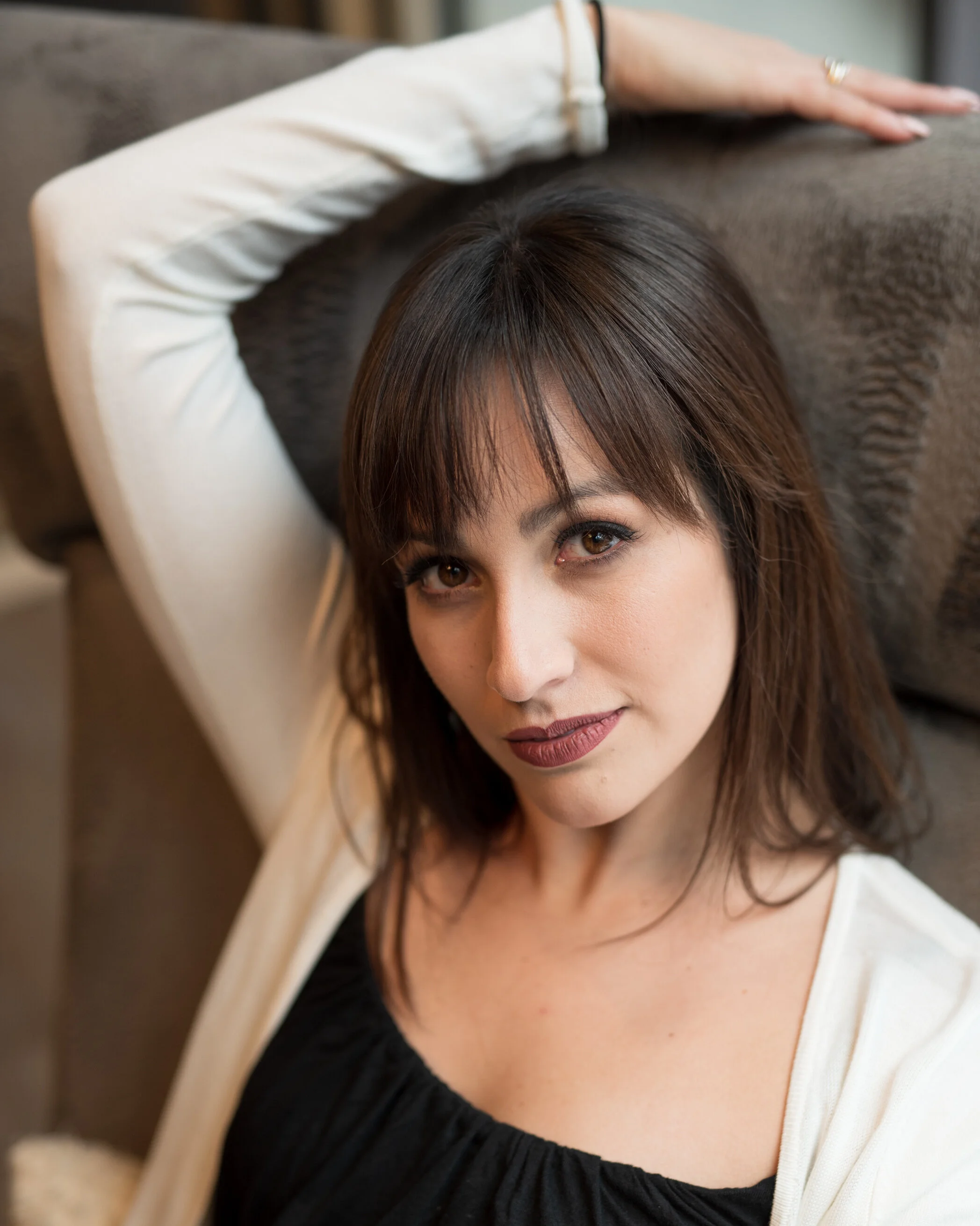Close-up of a woman with brown hair and makeup, posing with one arm resting on a headboard or furniture piece, looking directly at the camera.