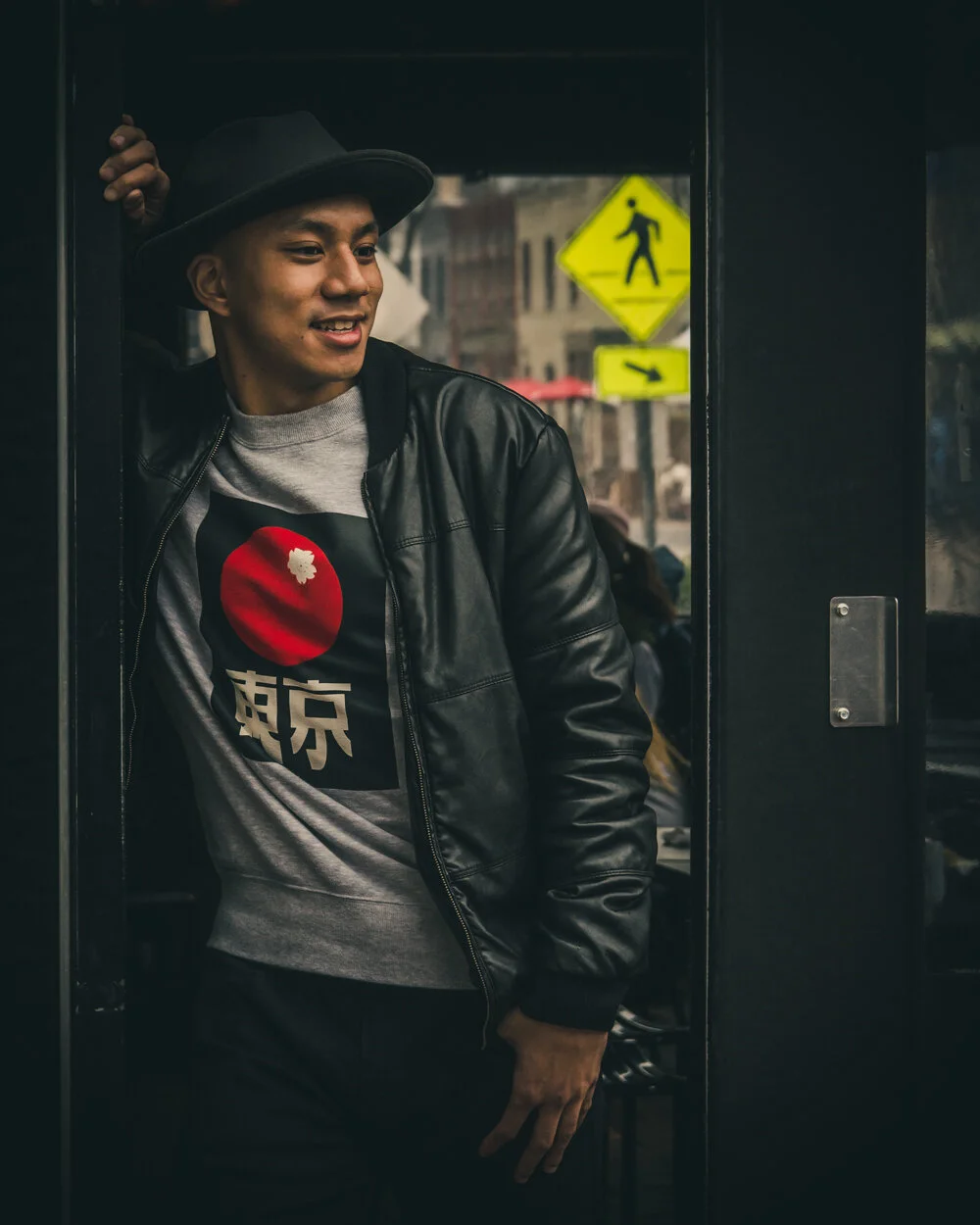 A young man wearing a grey shirt with a red Japanese flag and Japanese characters, a black leather jacket, and a grey fedora hat, leaning out of a door in an urban setting.