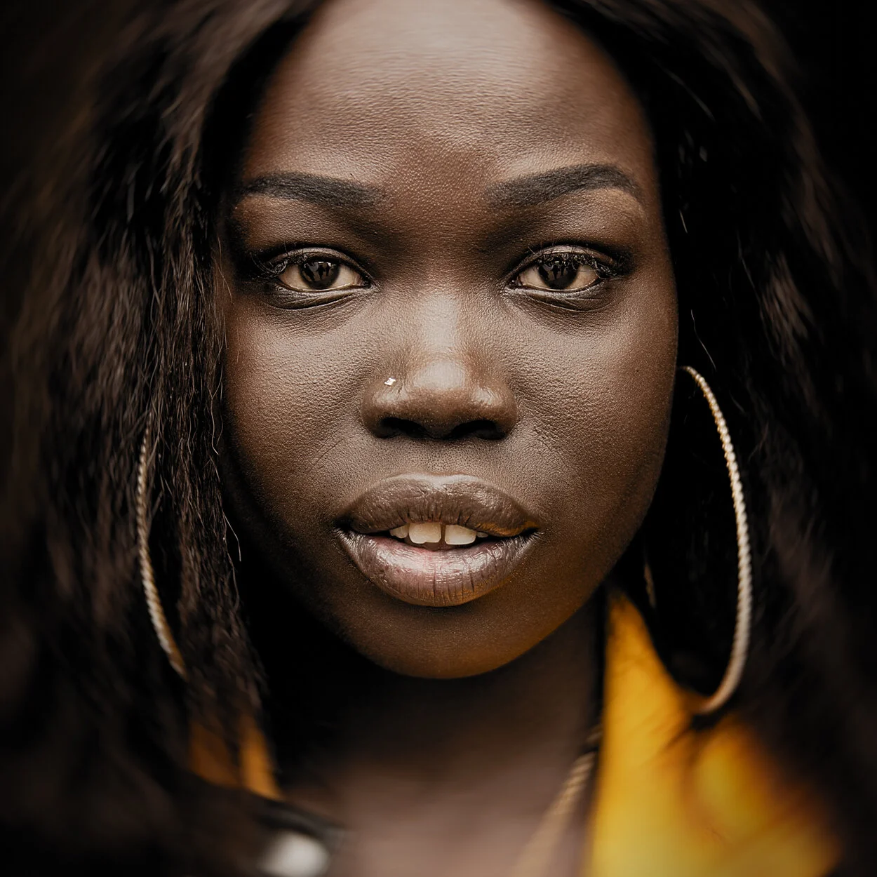 Close-up portrait of a Black woman with dark braided hair, gold hoop earrings, and a yellow garment, looking directly at the camera.