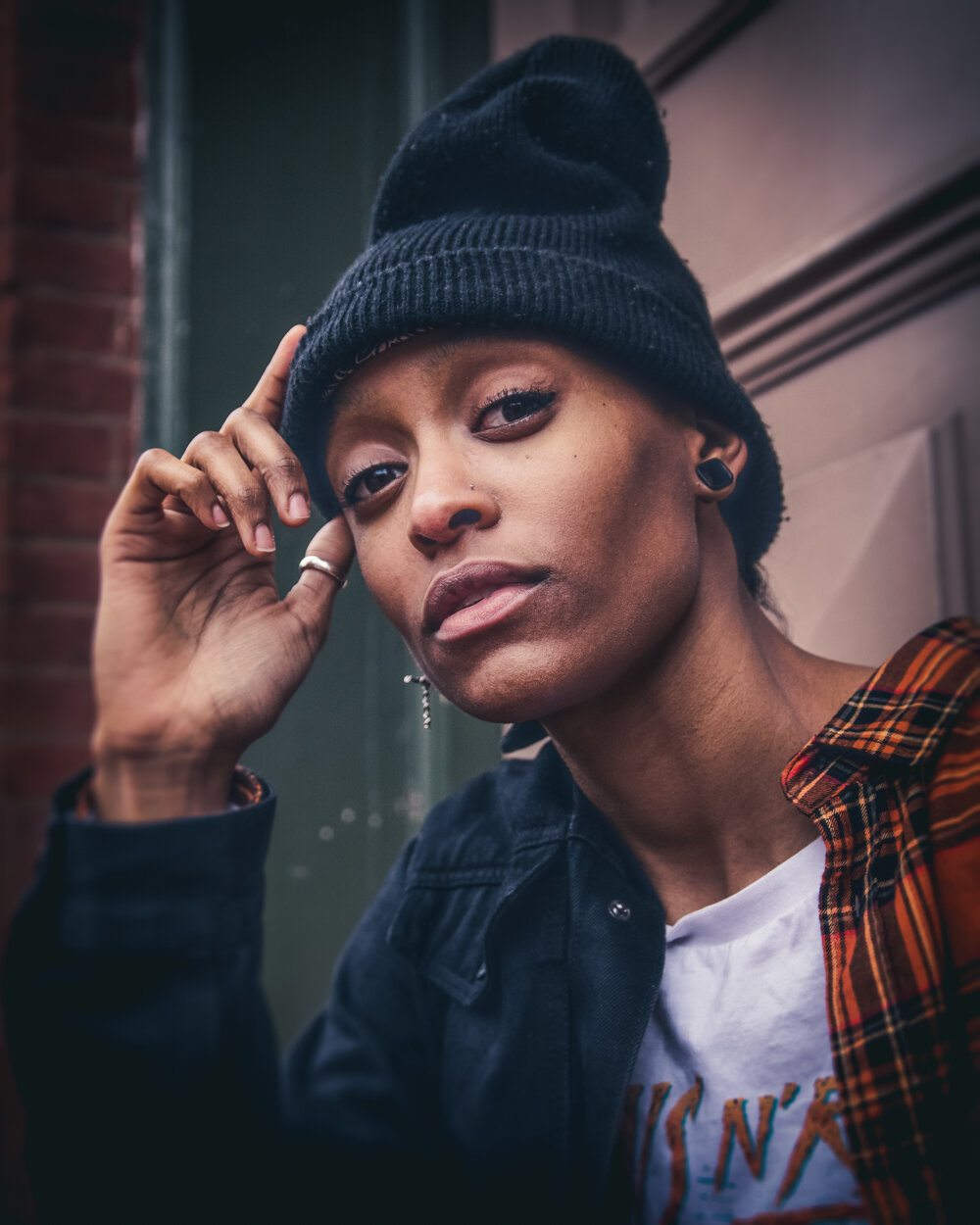 A young person with light skin, short hair, and multiple earrings, wearing a black beanie, a black denim jacket, and a white graphic t-shirt with an orange and black plaid shirt over it, resting their head on their hand outside near a brick wall and 