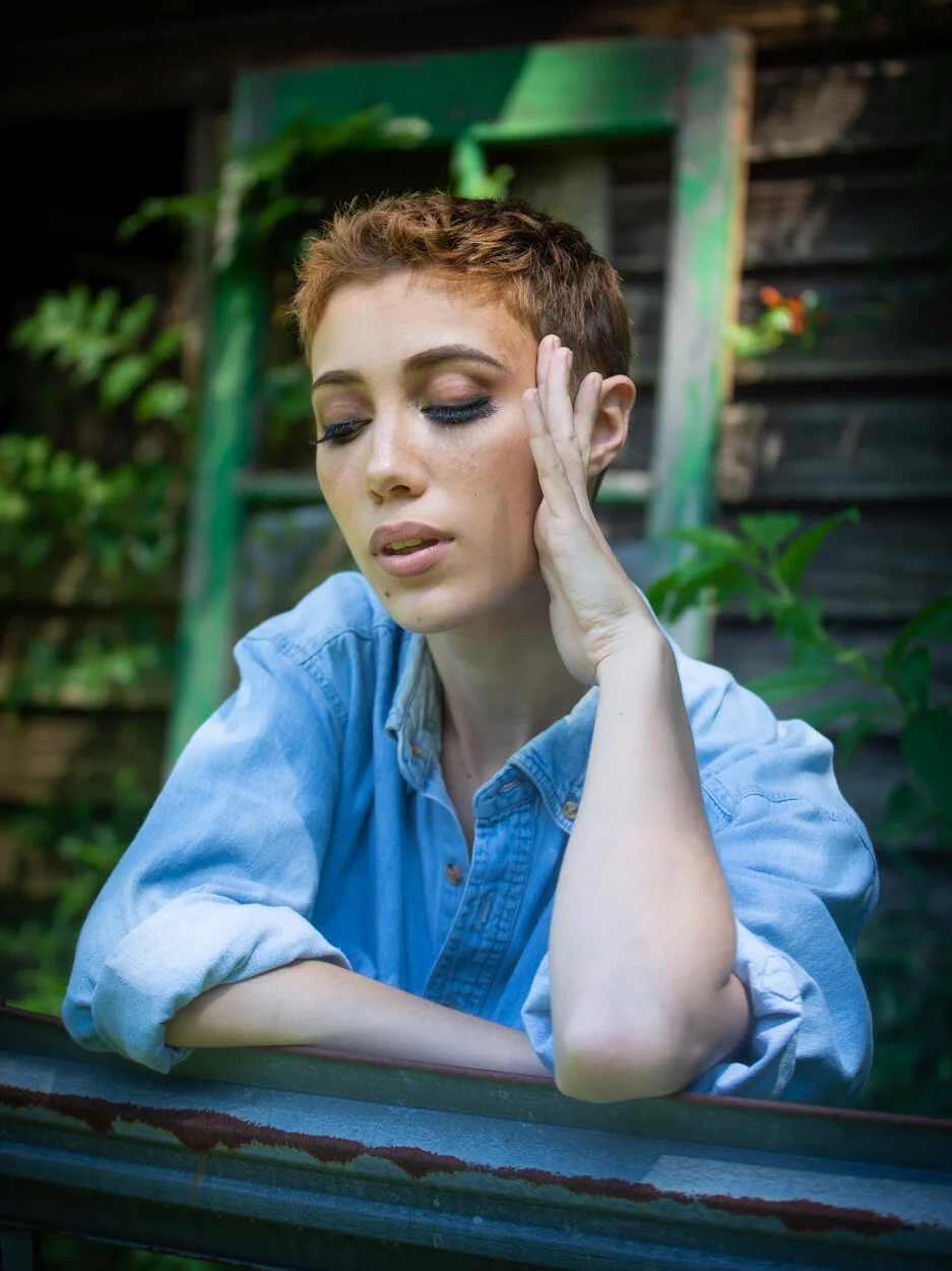 A woman with short red hair and makeup, resting her head on her hand with eyes closed, outdoors with greenery and a rustic wooden background.